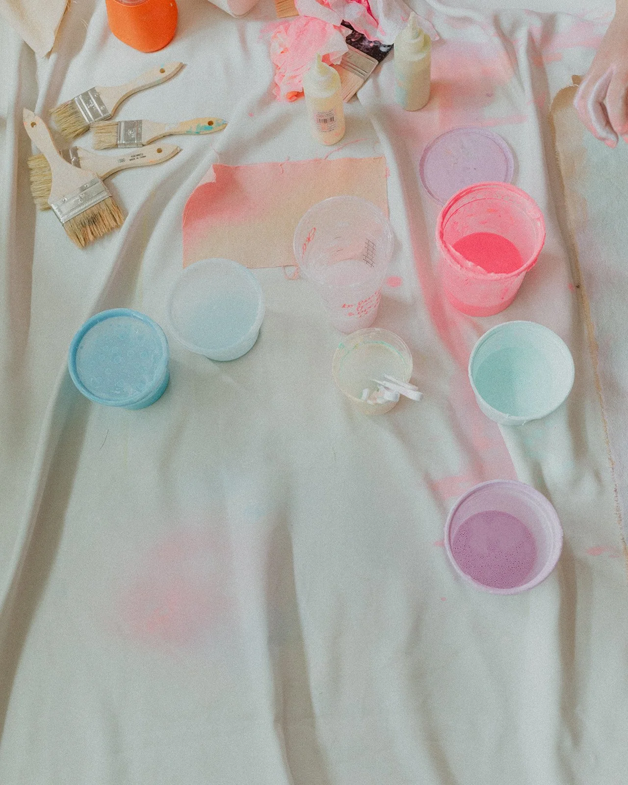 Paint supplies including brushes, containers of pastel pink, blue, green, and purple paint, paint bottles, on a painting drop cloth in a home studio.