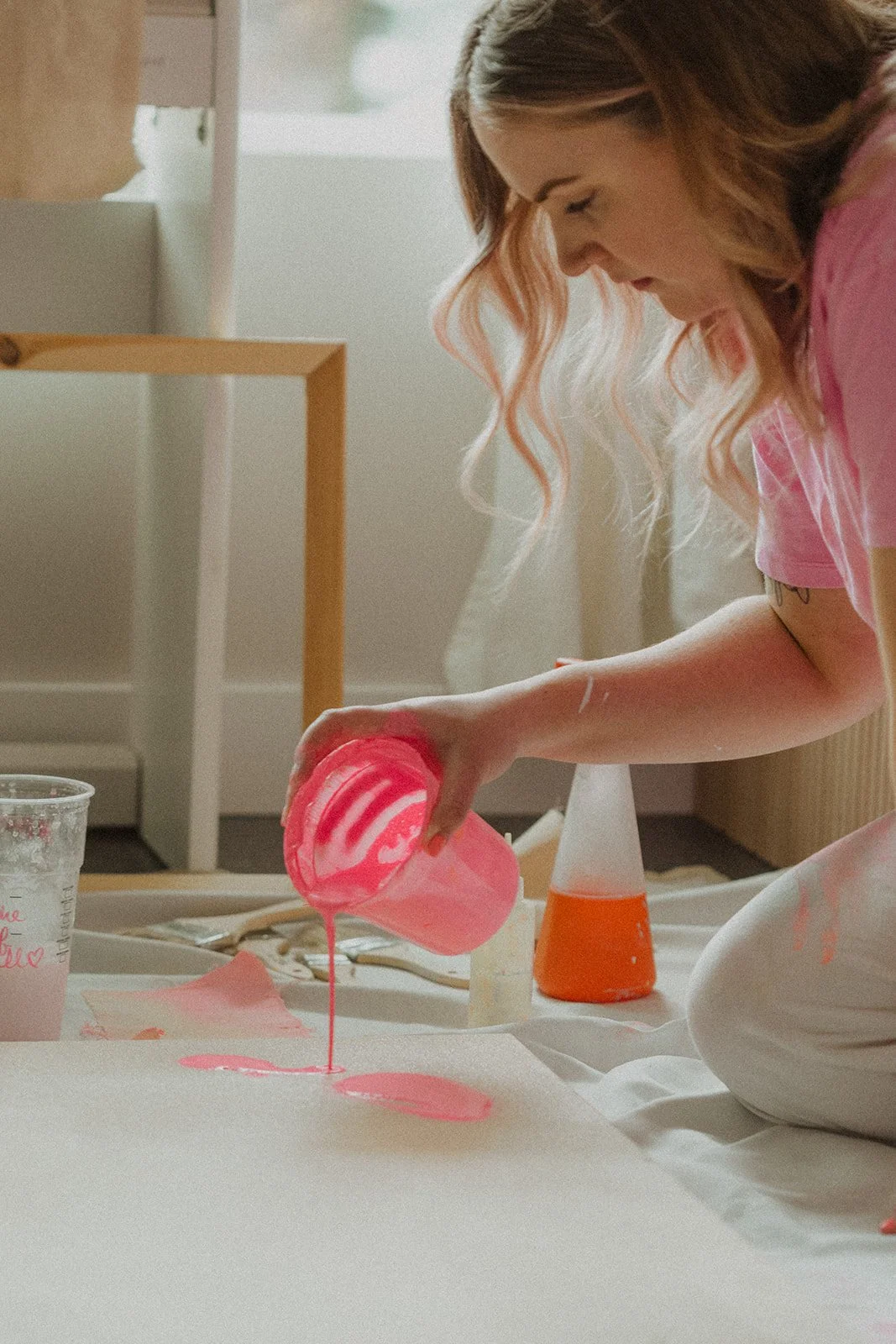 An artist with wavy pink hair pours pink paint onto a large canvas, with art supplies around her.