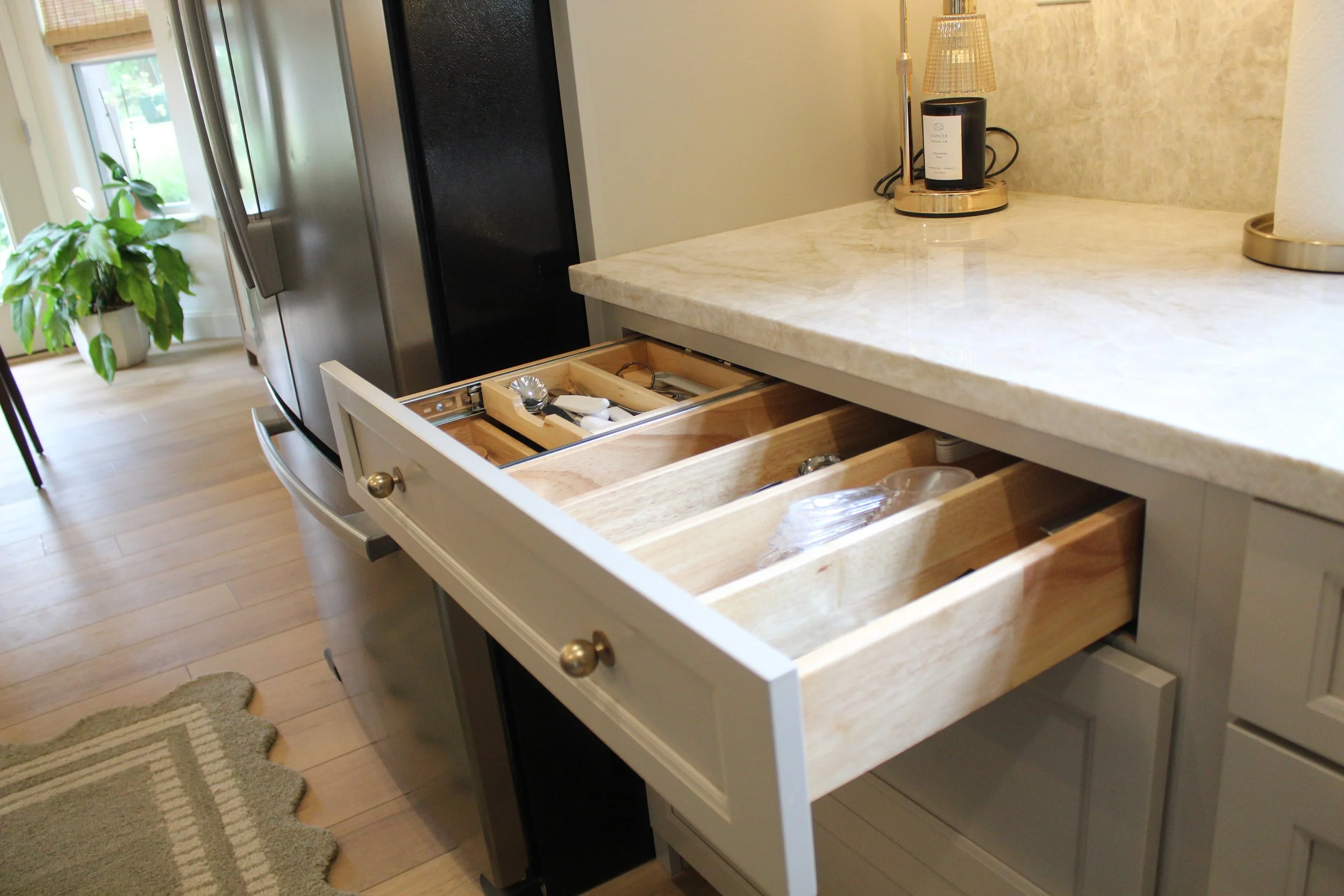 Kitchen with an open drawer containing small utensils and kitchen tools. A potted plant is on the windowsill, and a countertop with a lamp and paper towel roll.