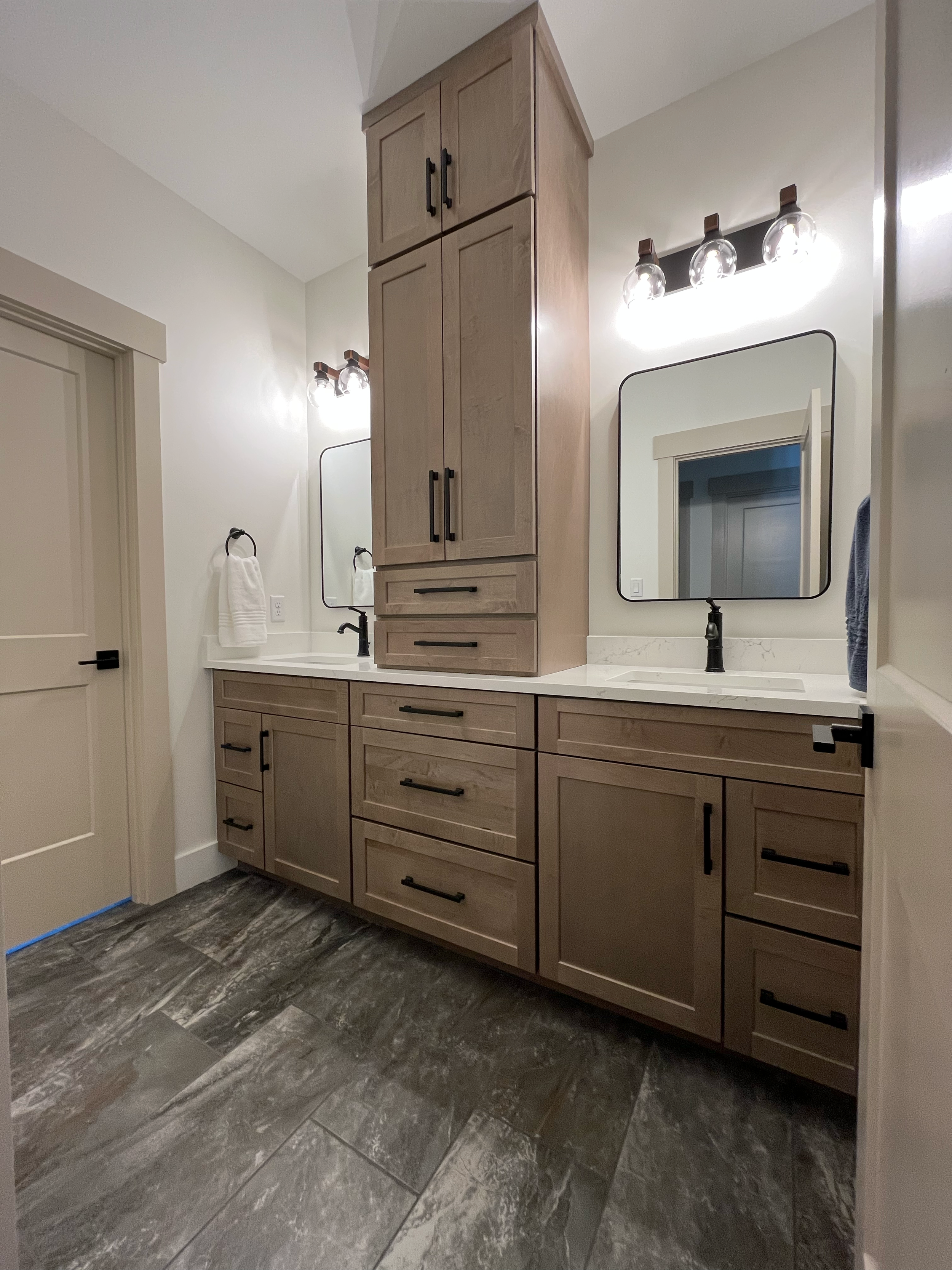 A modern bathroom vanity with wooden cabinets, black handles, and a marble countertop. There are two mirrors with black frames, black light fixtures with four bulbs each, and a small white sink. The floor has dark gray tile flooring.