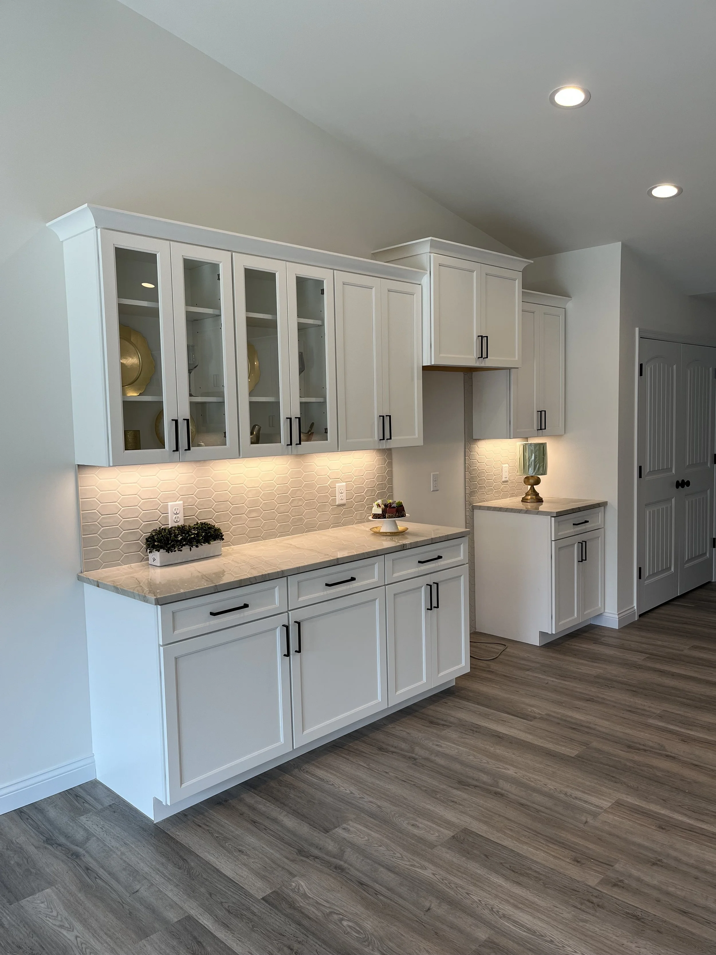 White kitchen cabinets with black handles, beige granite countertops, and a decorative backsplash with hexagonal tiles. The cabinets are above and below the counter, with some glass-front doors displaying dishes. There is a small golden lamp with a g