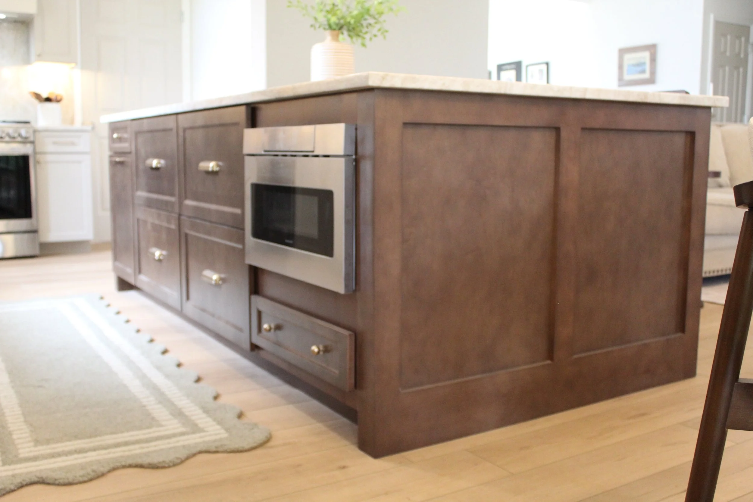 Kitchen island with wooden cabinets, drawers, and a built-in microwave oven, topped with a light-colored countertop, in a bright kitchen with hardwood flooring.