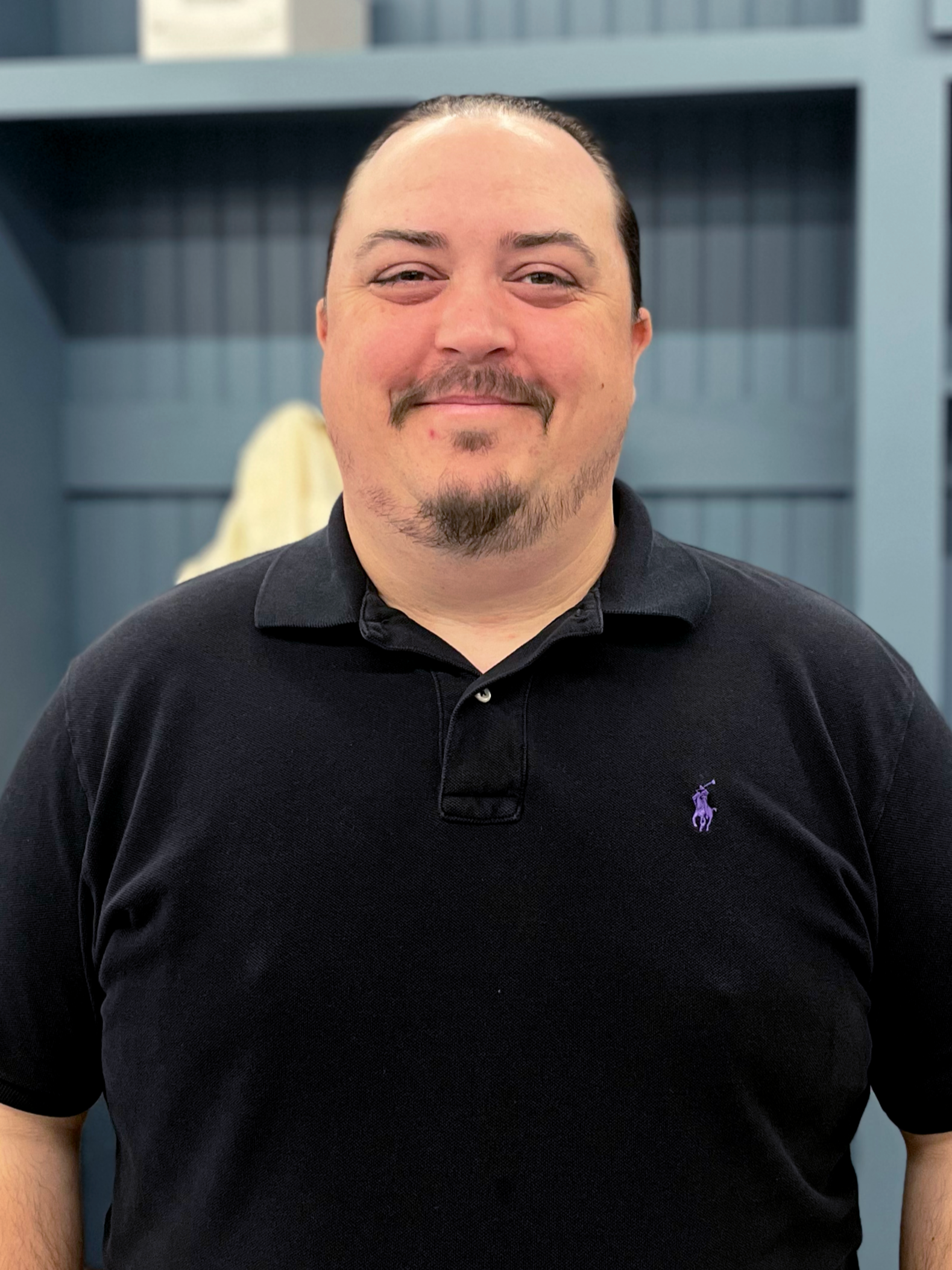 Close-up of a smiling man with a goatee and mustache, wearing a black polo shirt with a purple logo, standing in front of a blue background with shelves.