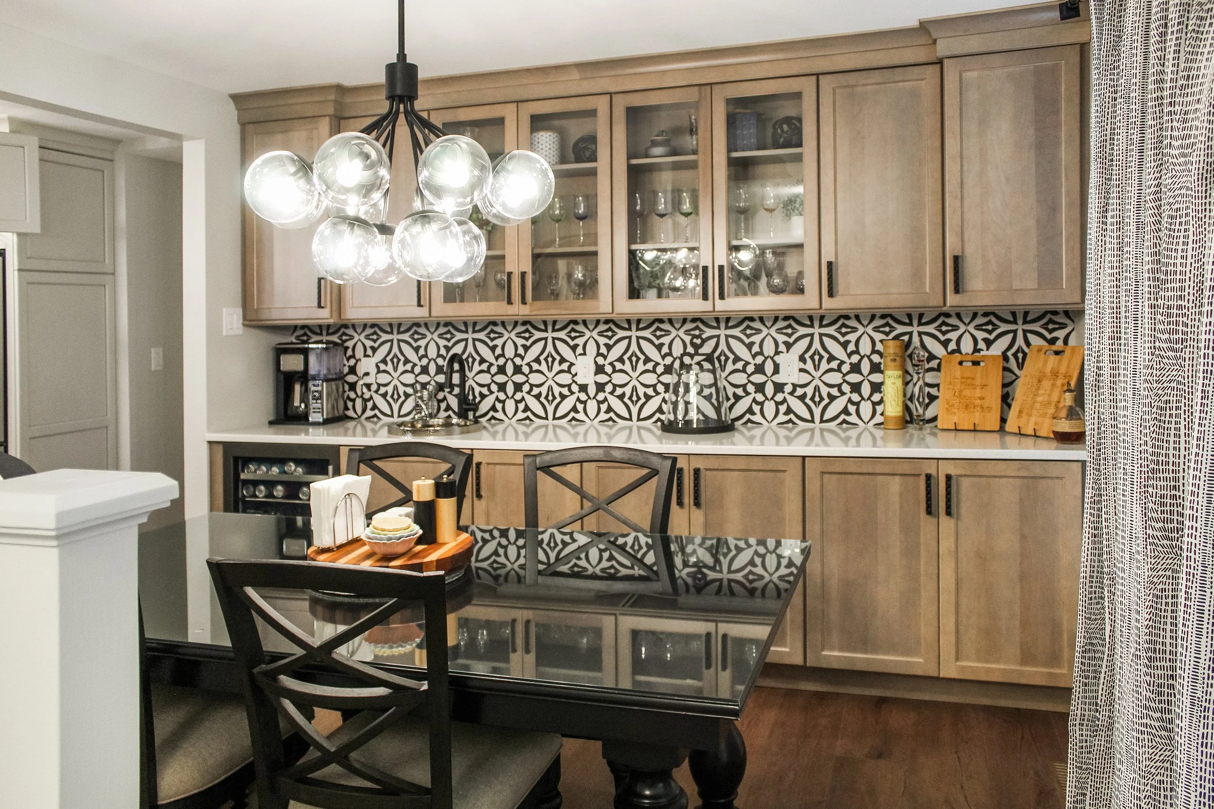 A modern kitchen with wooden upper cabinets, black and white patterned backsplash, black countertops, a glass-top dining table, black chairs, and a distinctive chandelier with multiple glass globes.