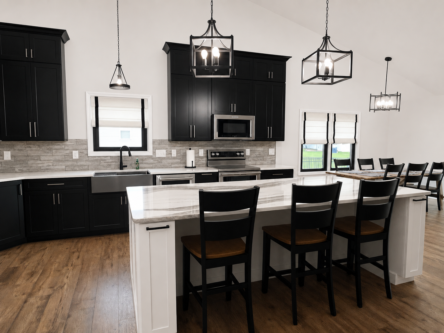 Modern kitchen with black cabinets, white marble countertops, stainless steel appliances, a kitchen island with four black chairs, pendant lighting, and windows with white shutters.