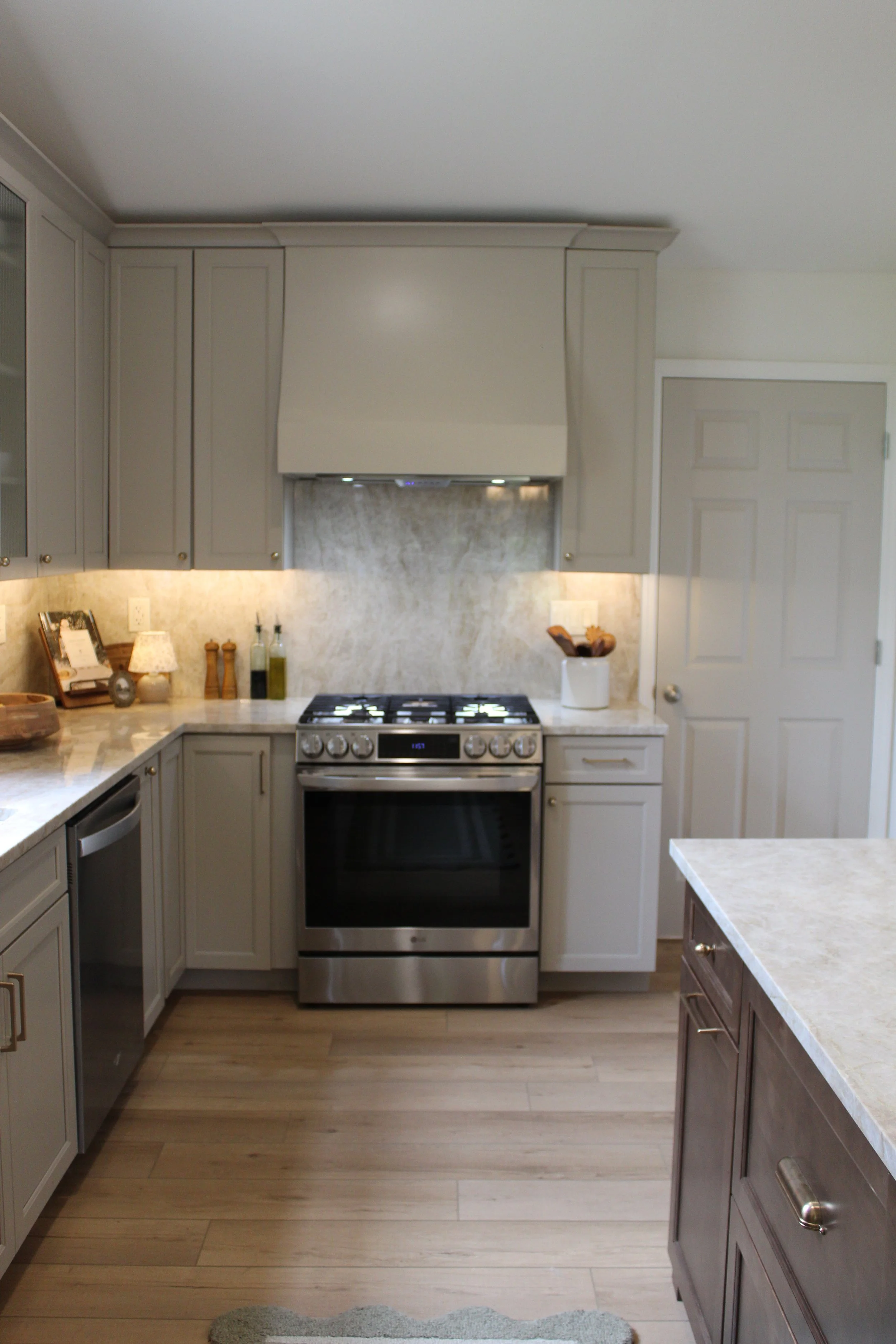 A modern kitchen with light gray cabinetry, a stainless steel stove, marble countertops, and light wood flooring.