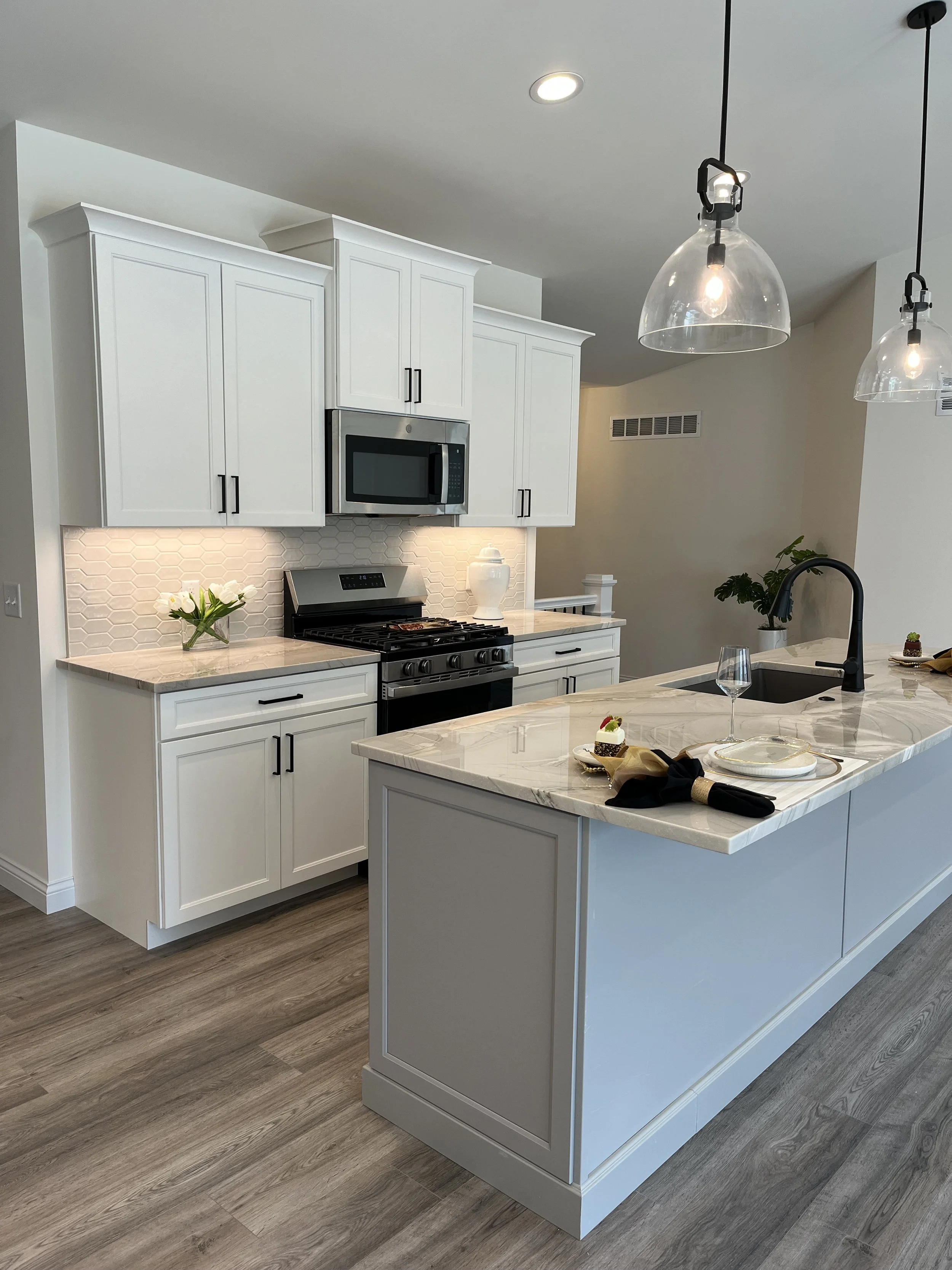 Modern kitchen with white cabinets, stainless steel microwave and stove, hexagonal tile backsplash, marble countertop, kitchen island with a sink, some plates, a glass of wine, and a small cake, pendant lights, and a wooden floor.