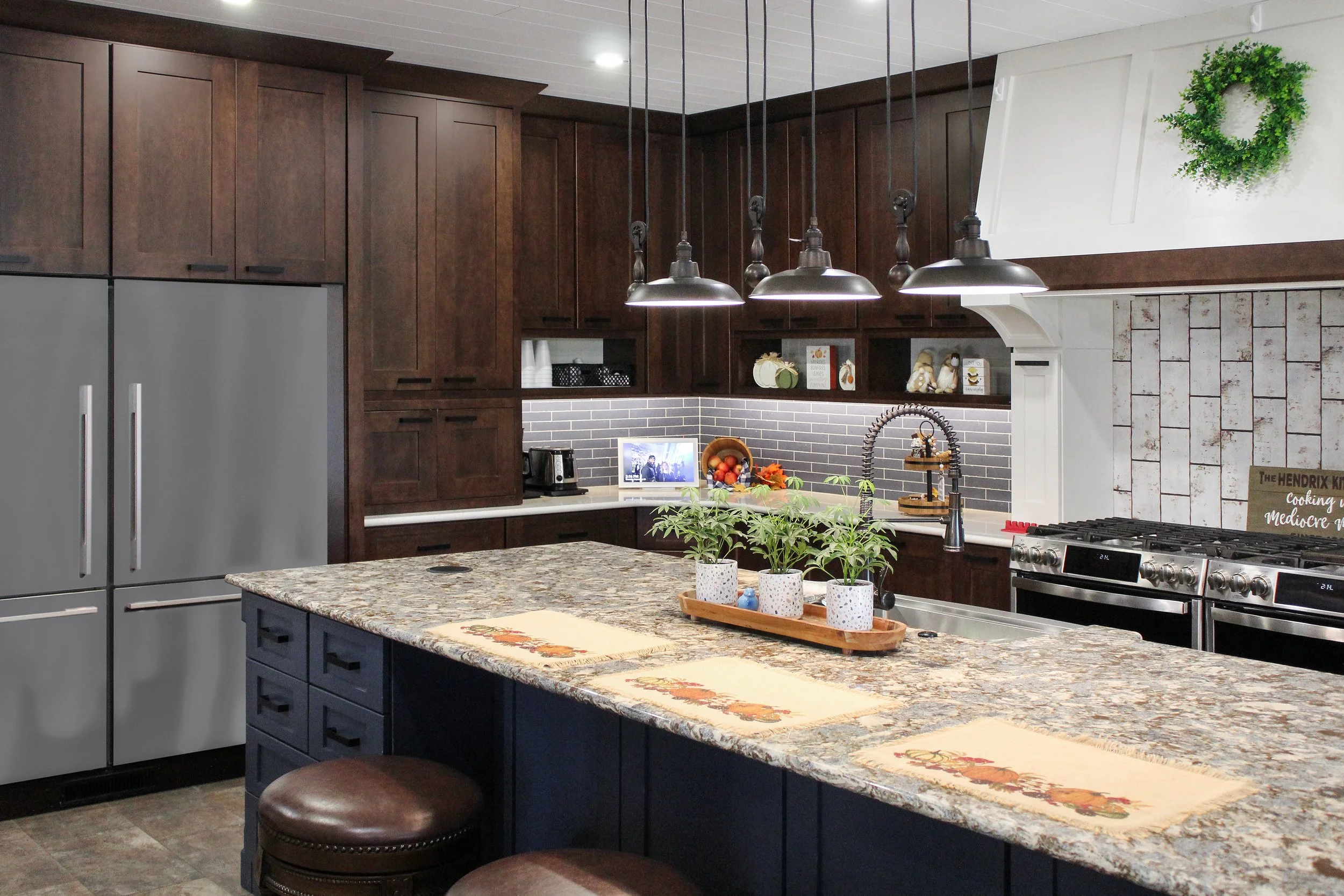 A modern kitchen with dark wooden cabinets, a granite countertop island with navy blue cabinets, and stainless steel appliances including a refrigerator and a stove. The kitchen features pendant lights, potted herbs, a small digital photo frame, and 