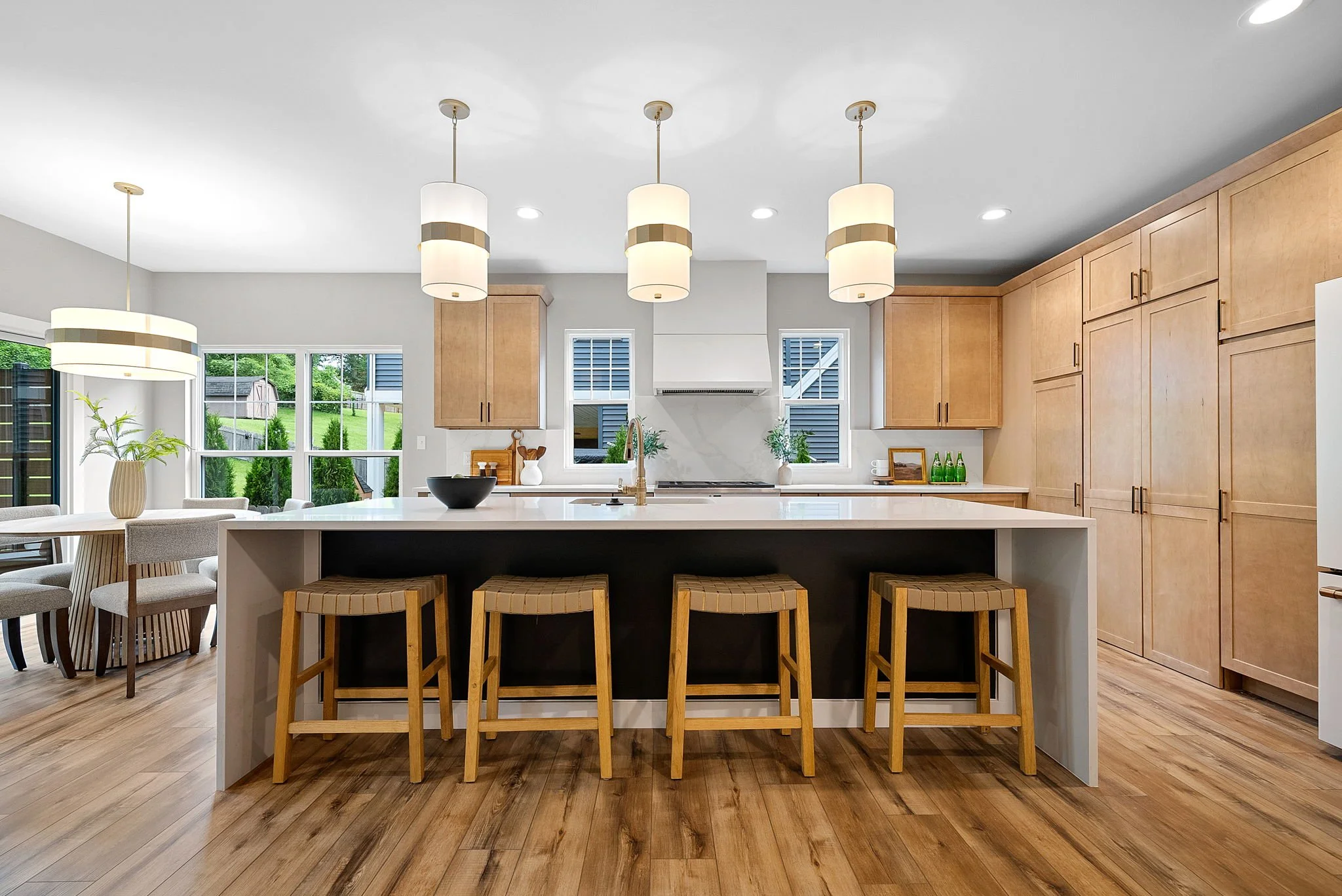 Modern kitchen with wooden cabinets, island with four stools, pendant lights, and large windows with outdoor view.