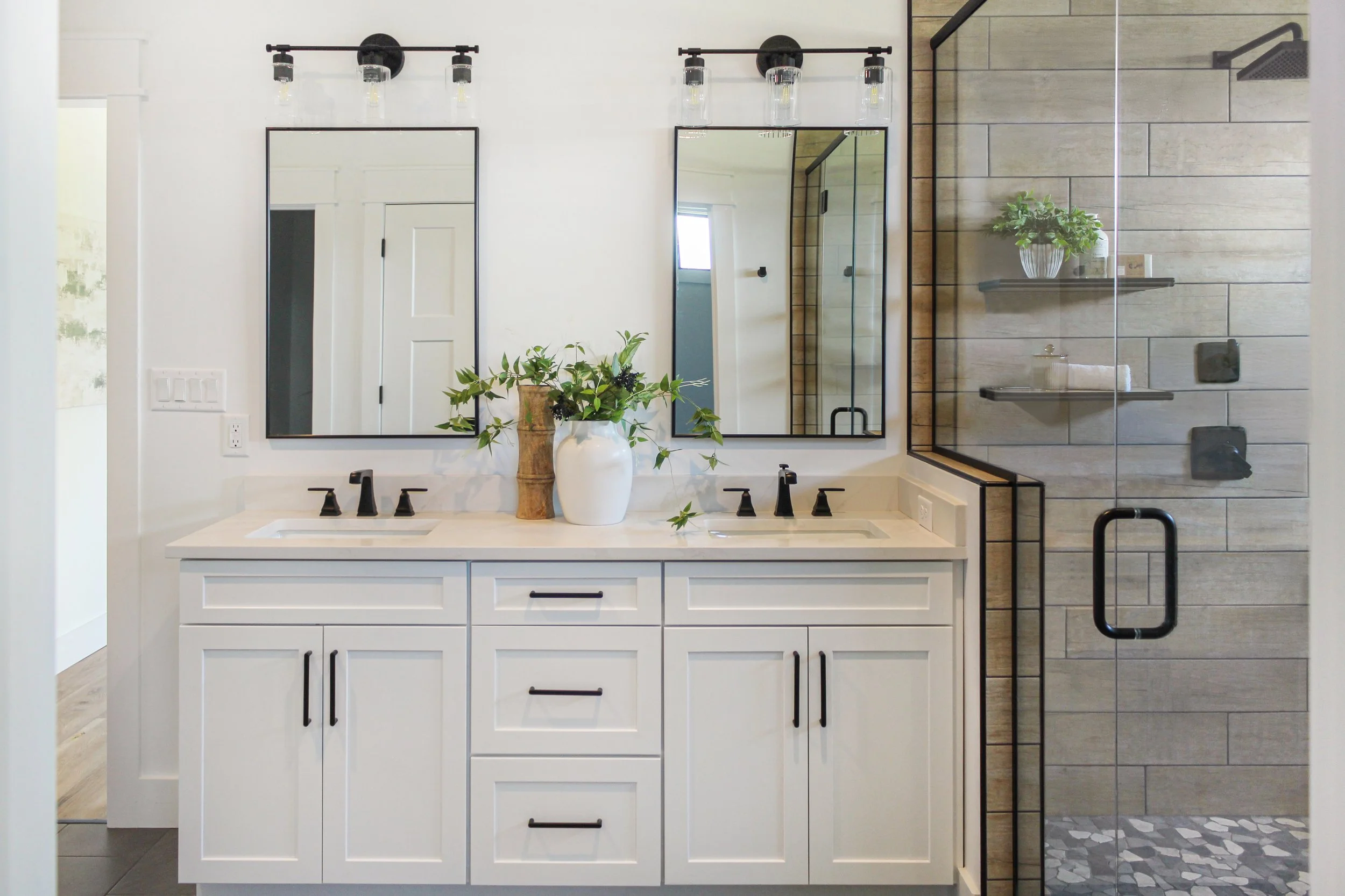 Modern bathroom with double vanity, large mirrors, black fixtures, and a glass-enclosed walk-in shower with beige tile and decorative shelves.