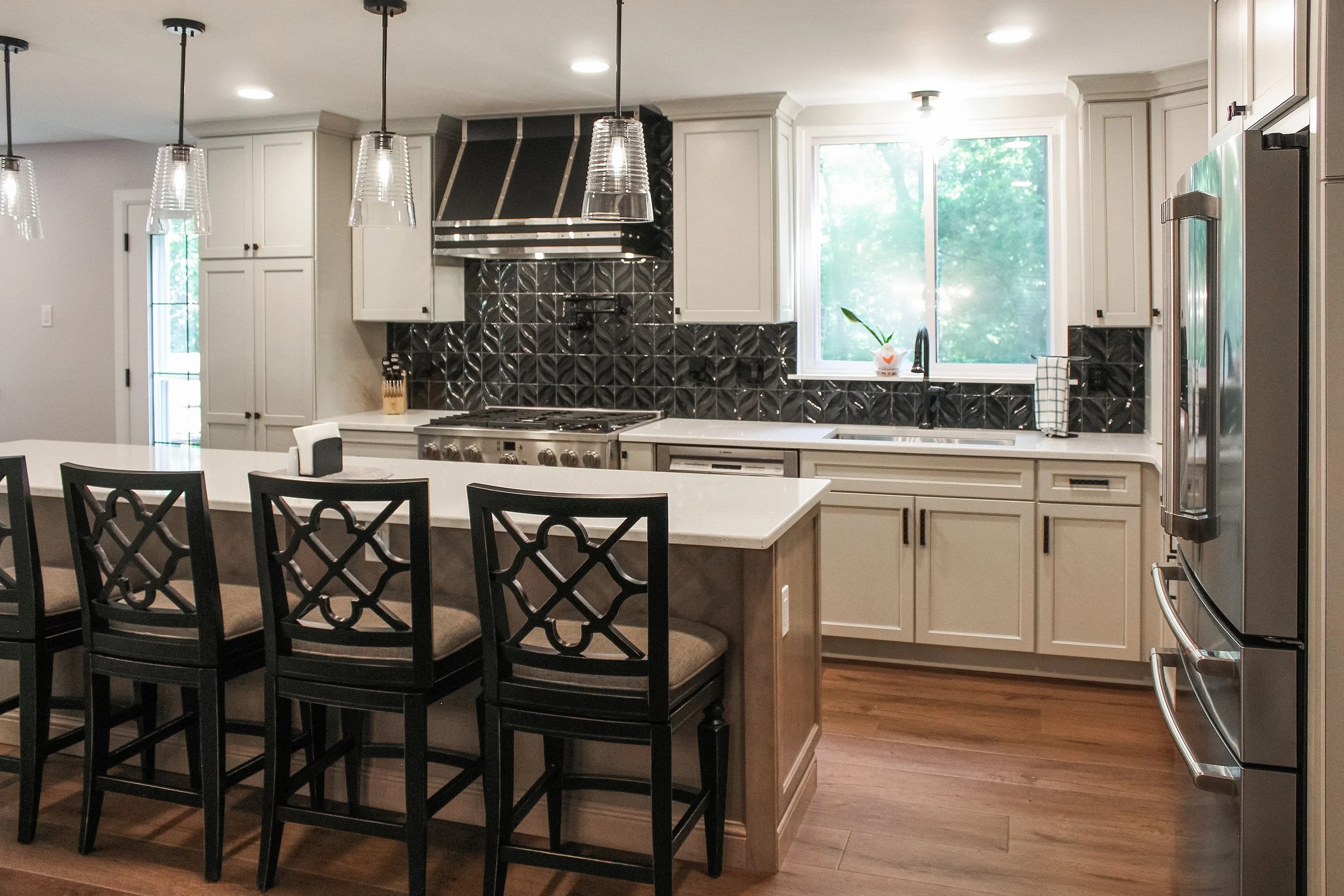 Modern kitchen with white cabinets, black geometric backsplash, stainless steel appliances, a window above the sink, and a breakfast bar with four black chairs.