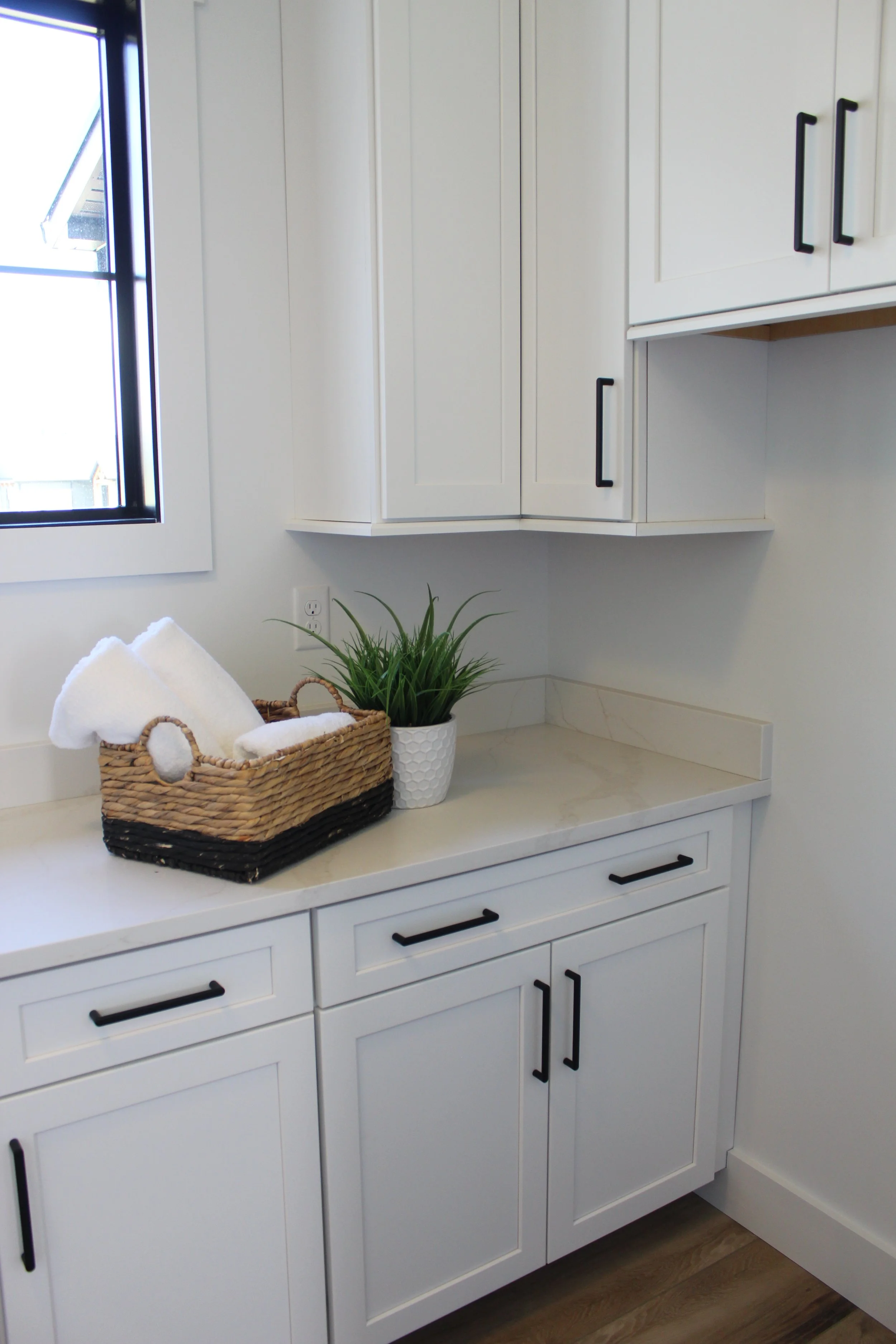 White kitchen with cabinets, black handles, a window, and a countertop with a basket of rolled white towels and a potted plant.