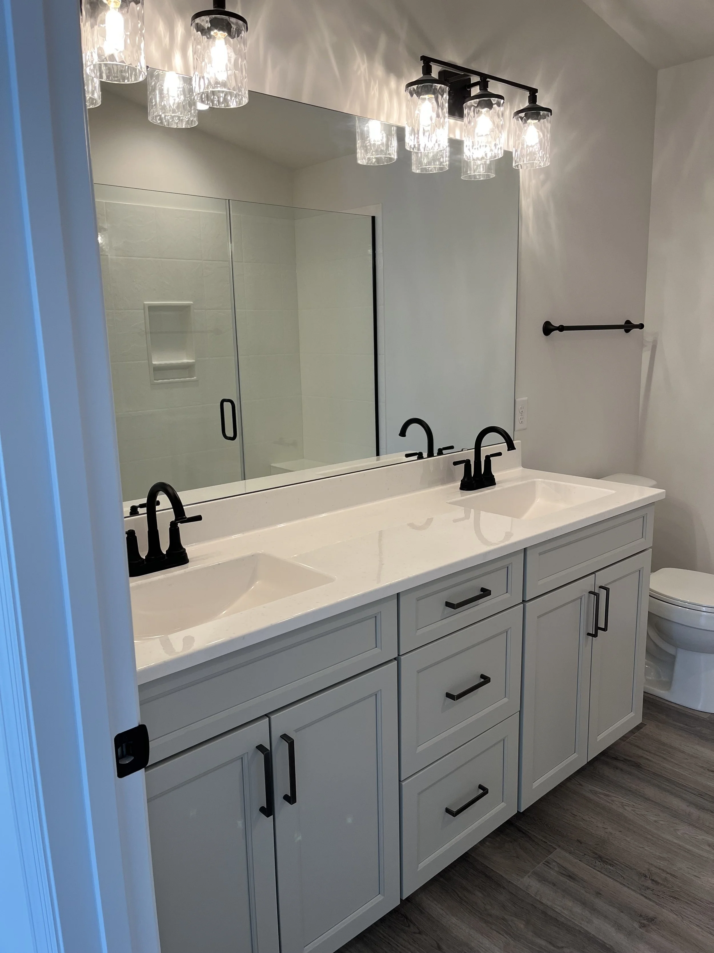 Bathroom with a double vanity, white cabinets, black hardware, large mirror, black light fixtures, and a shower with glass doors.