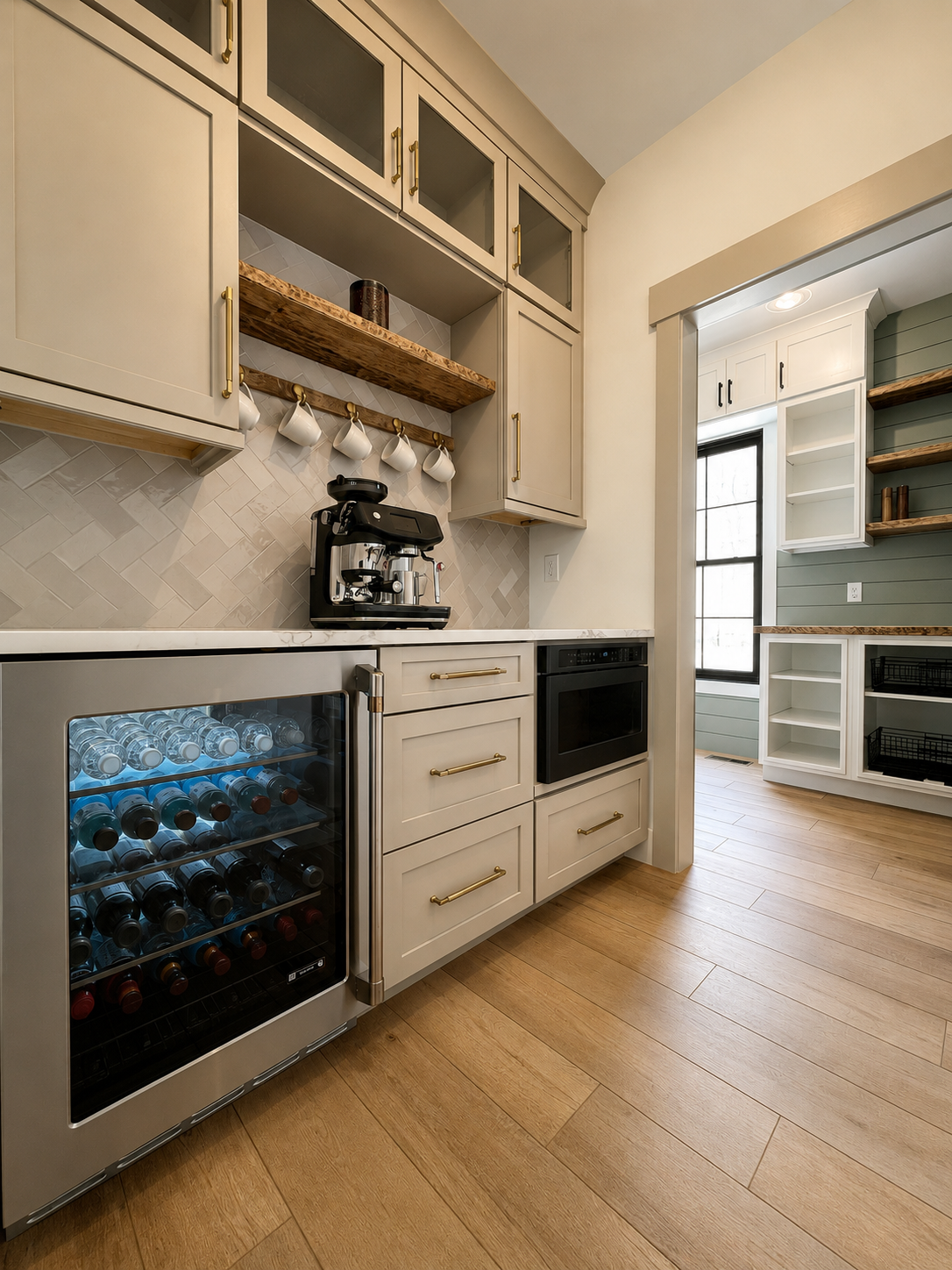 Kitchen with beige cabinets, a built-in wine cooler stocked with water bottles, a black coffee machine, and an oven. Open shelving with wooden and white shelves, a window with natural light, and wooden flooring.