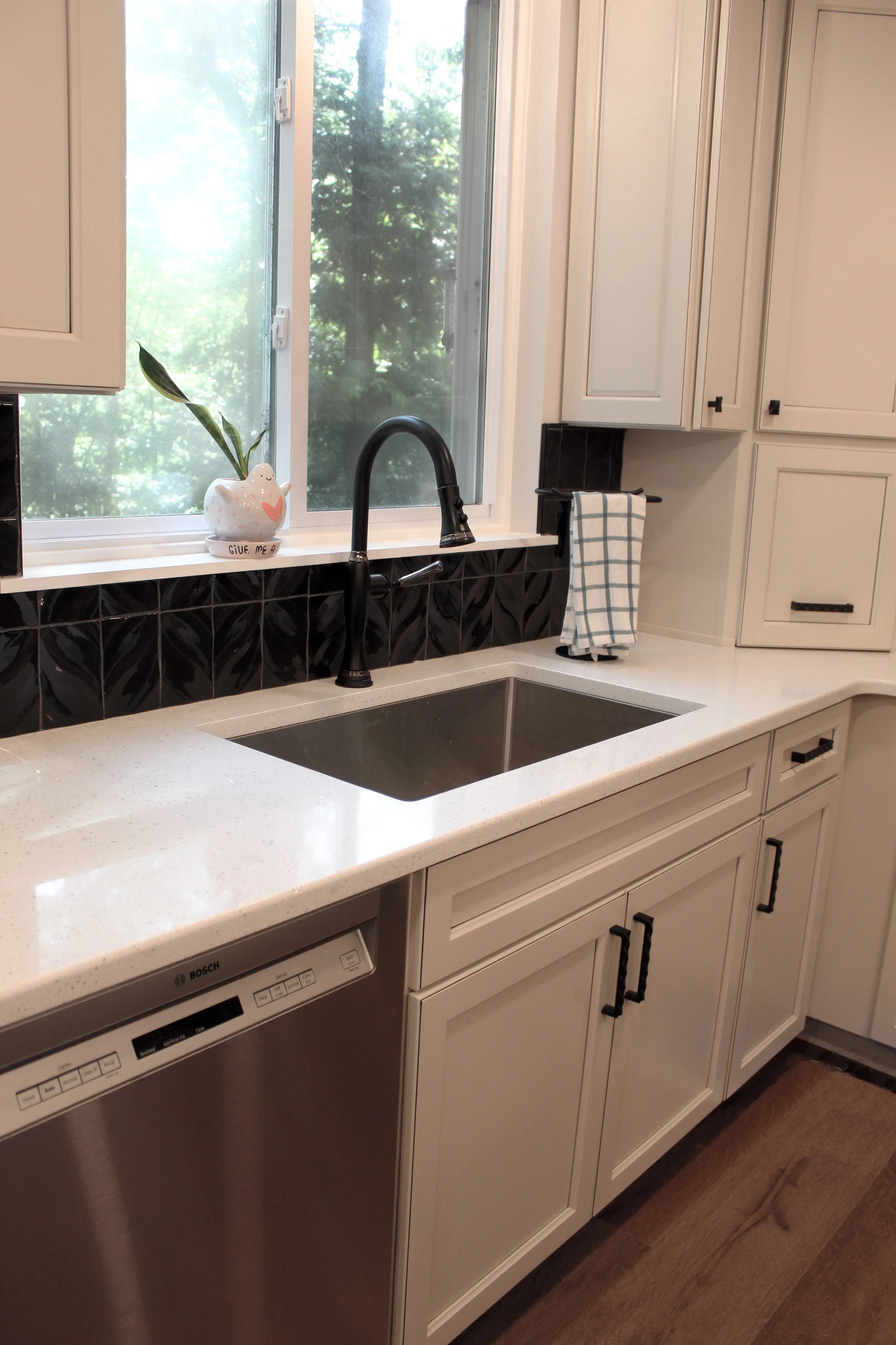 A kitchen with white cabinetry, a black faucet and sink, a window with greenery outside, a small vase with a plant, and a dishwasher.