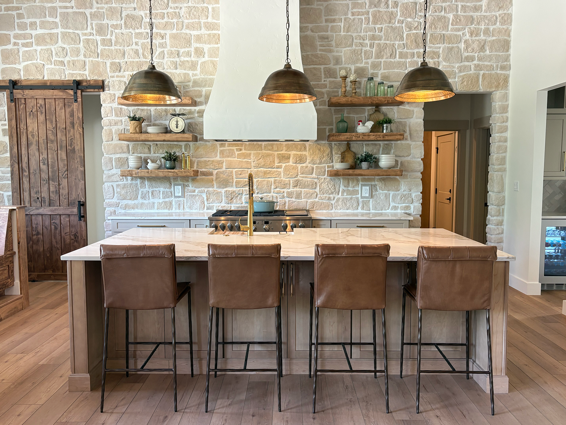 The image shows a modern kitchen with a large marble island counter and five brown leather chairs. Behind the counter, there is a white brick wall with open wooden shelves displaying various decor items, including bowls, vases, and small potted plant