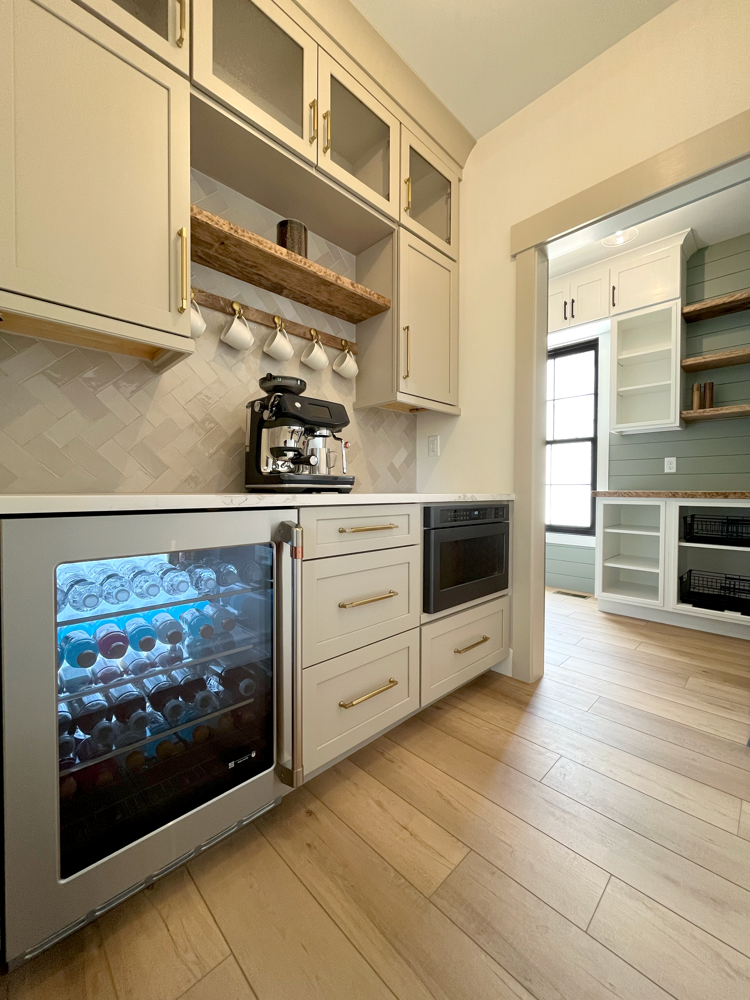 A kitchen corner with cream cabinets, a wine cooler filled with bottled water, a coffee machine on the countertop, open shelves, and a window allowing natural light.