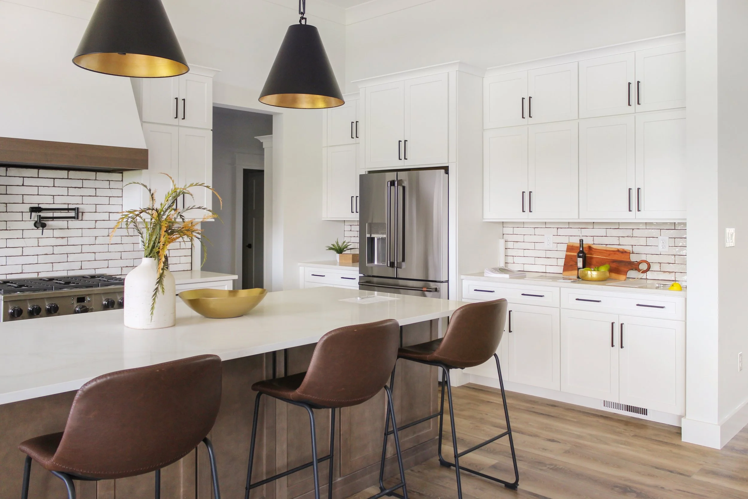 Modern kitchen with white cabinets, stainless steel refrigerator, black and gold pendant lights, and a kitchen island with brown leather chairs. Decor includes a white vase with plants and a yellow bowl.