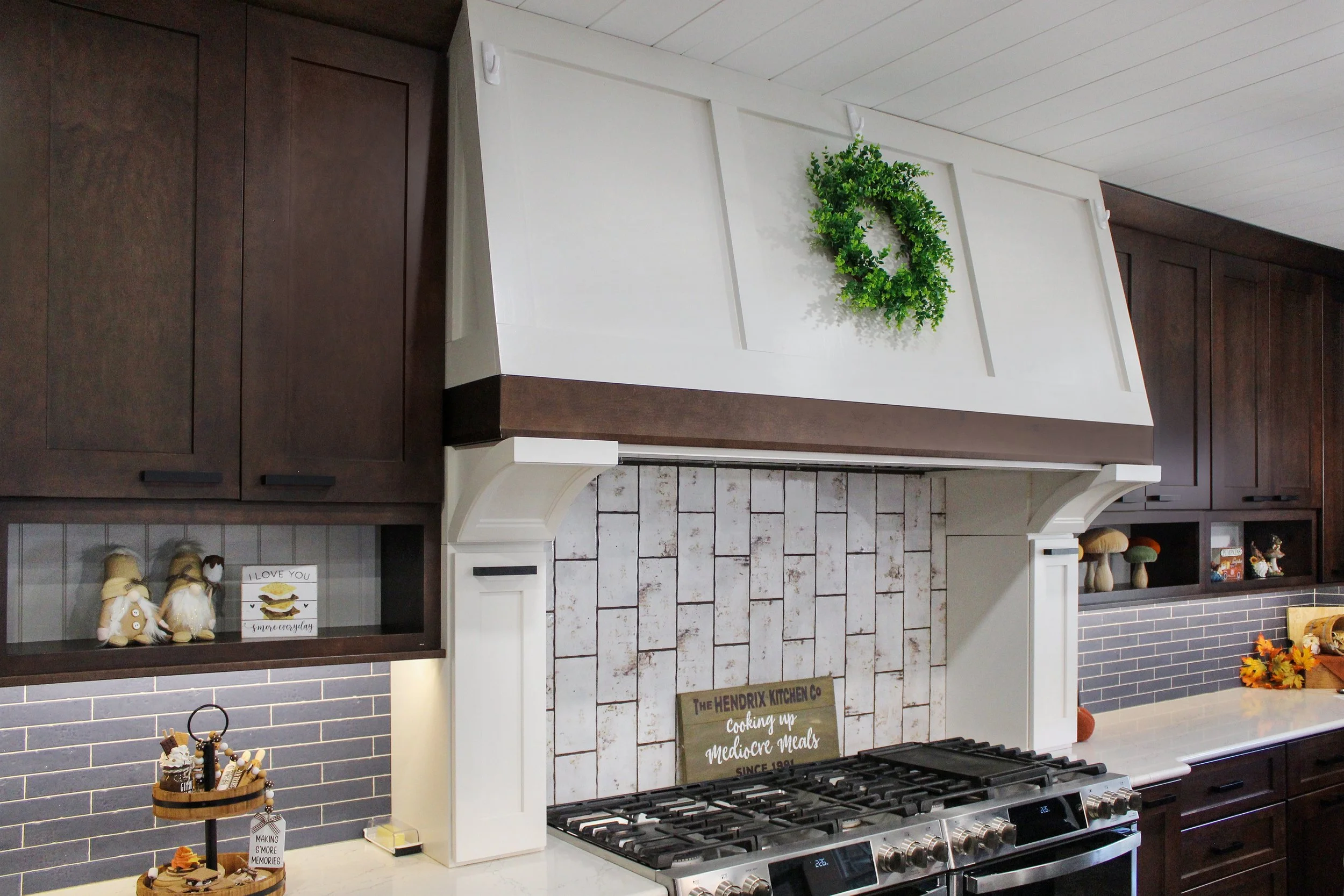 Kitchen with white and dark wood cabinets, a stove, decorative shelves, a sign, and a wreath on the vent hood.