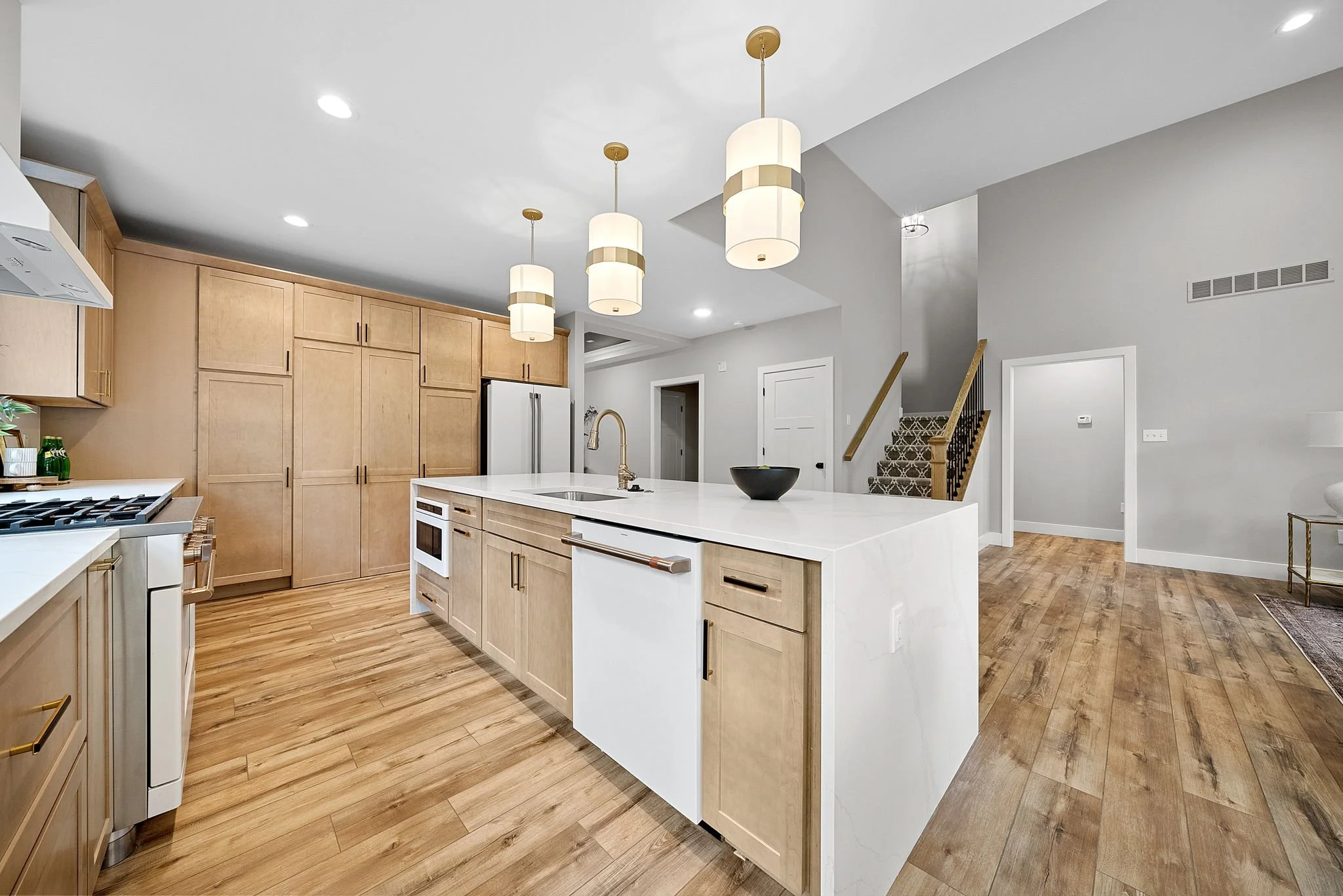 Modern kitchen with light wood cabinets, white countertops, and a white island with a black bowl, featuring three pendant lights above and hardwood floors.