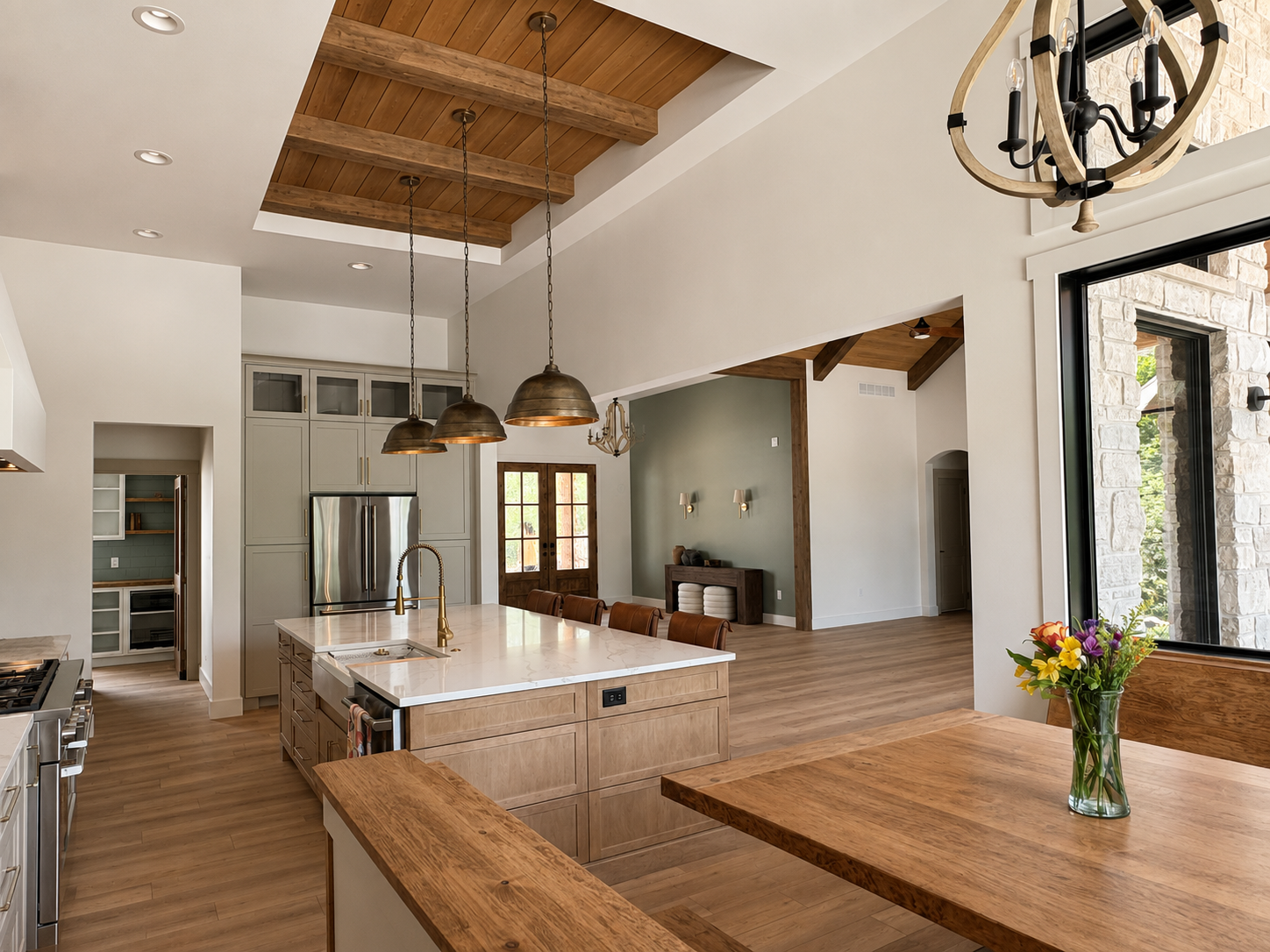 Open-concept kitchen with a large island, pendant lighting, stainless steel appliances, wooden beams on the ceiling, a dining table with a vase of colorful flowers, and a view into the living area with wood accents and a front door in the background.