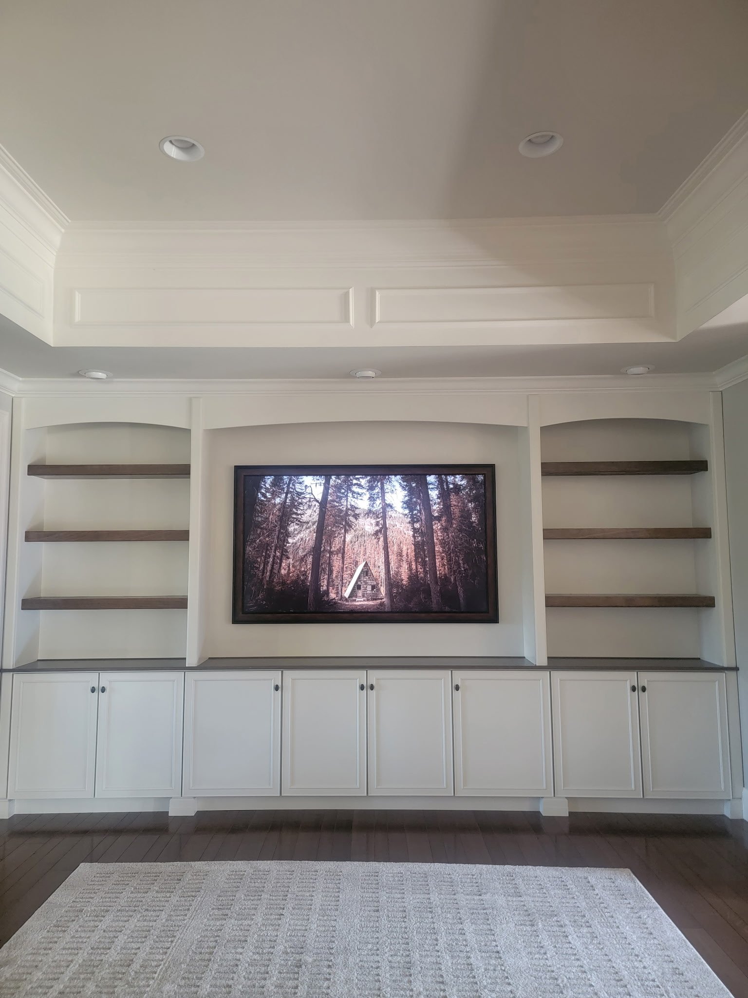 Living room with built-in white cabinets and open shelves, a large window with a forest view, and a flat-screen TV in the center.