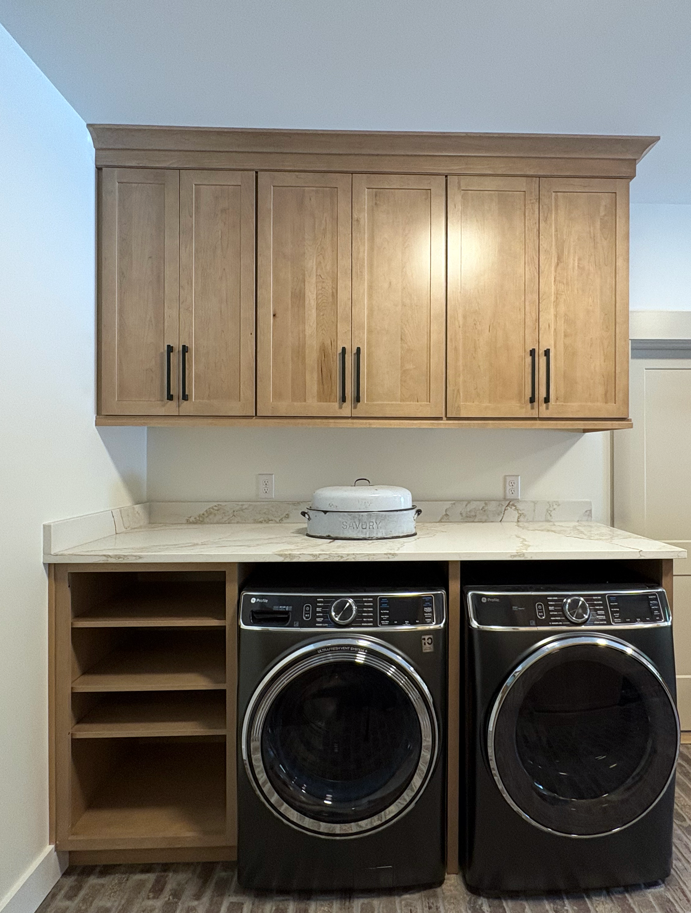 A laundry room with wooden cabinets above a countertop, a washing machine and dryer side by side, and empty shelves on the left. A round metal container with the word 'SAVORY' on it is placed on the countertop.