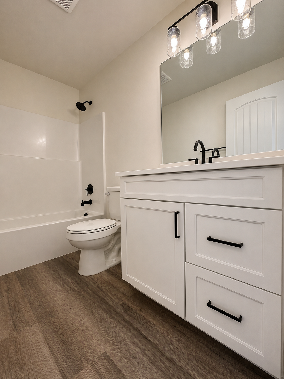 Bathroom with white vanity, black hardware, large mirror, wall-mounted light fixture, toilet, and bathtub with black fixtures, wood-look flooring.