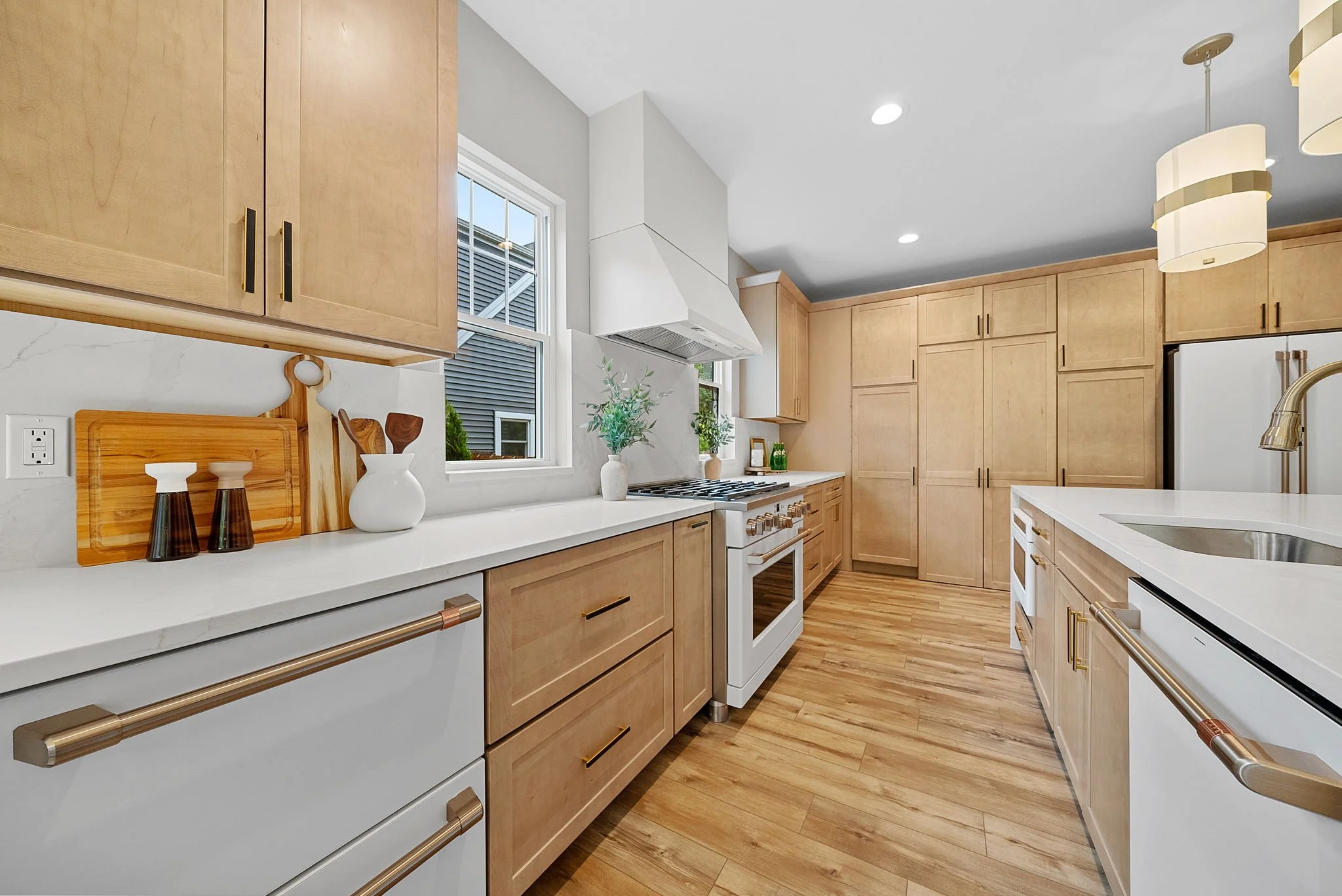 Modern kitchen with light wood cabinets, white countertops, a stainless steel refrigerator, and a white range hood. There are potted plants, decorative items, and a window letting in natural light. Wooden flooring complements the neutral color scheme