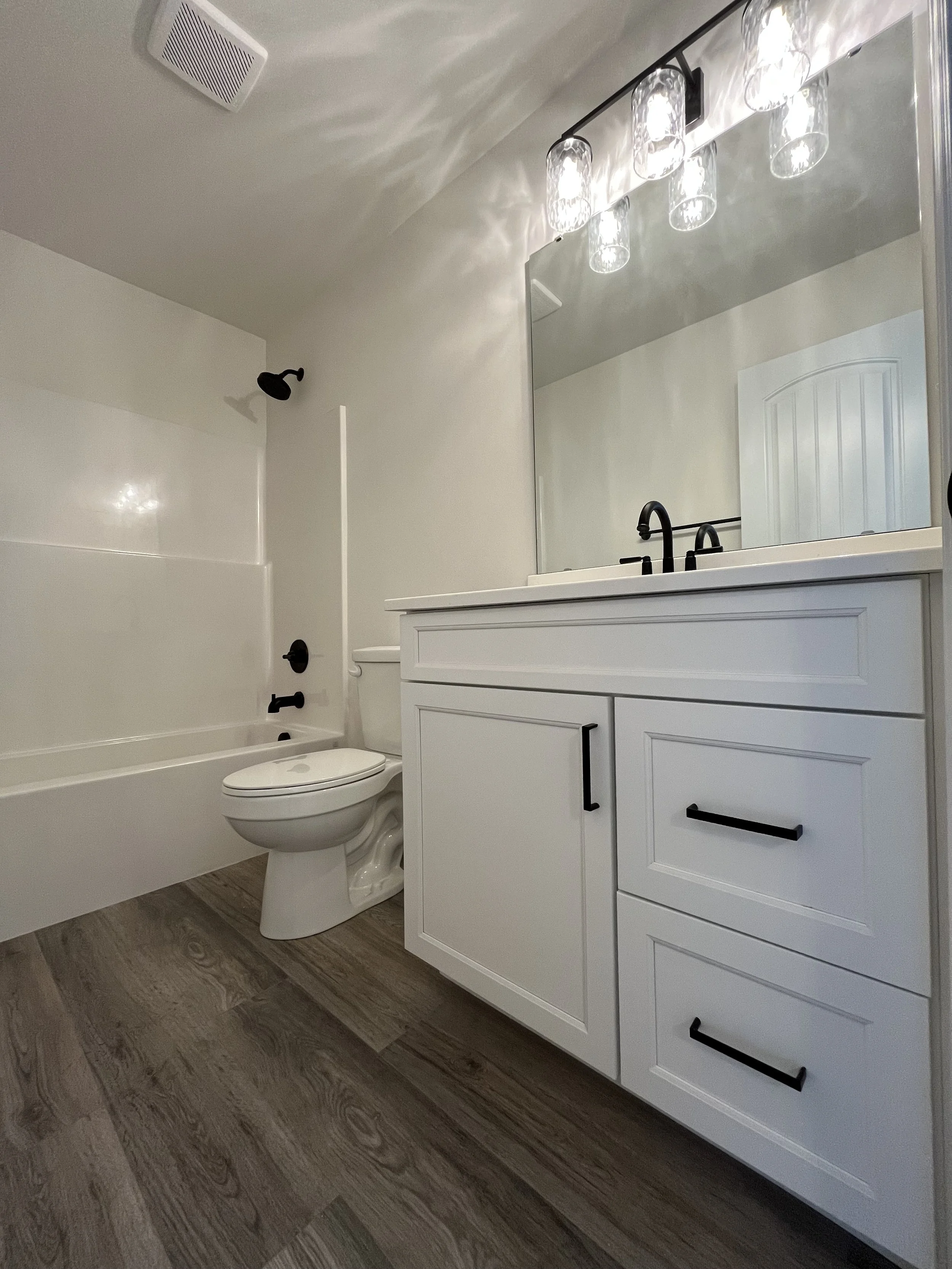 Bathroom with a white vanity cabinet, a large mirror, black faucet, overhead lighting fixture, toilet, shower with black fixtures, and wood-textured flooring.