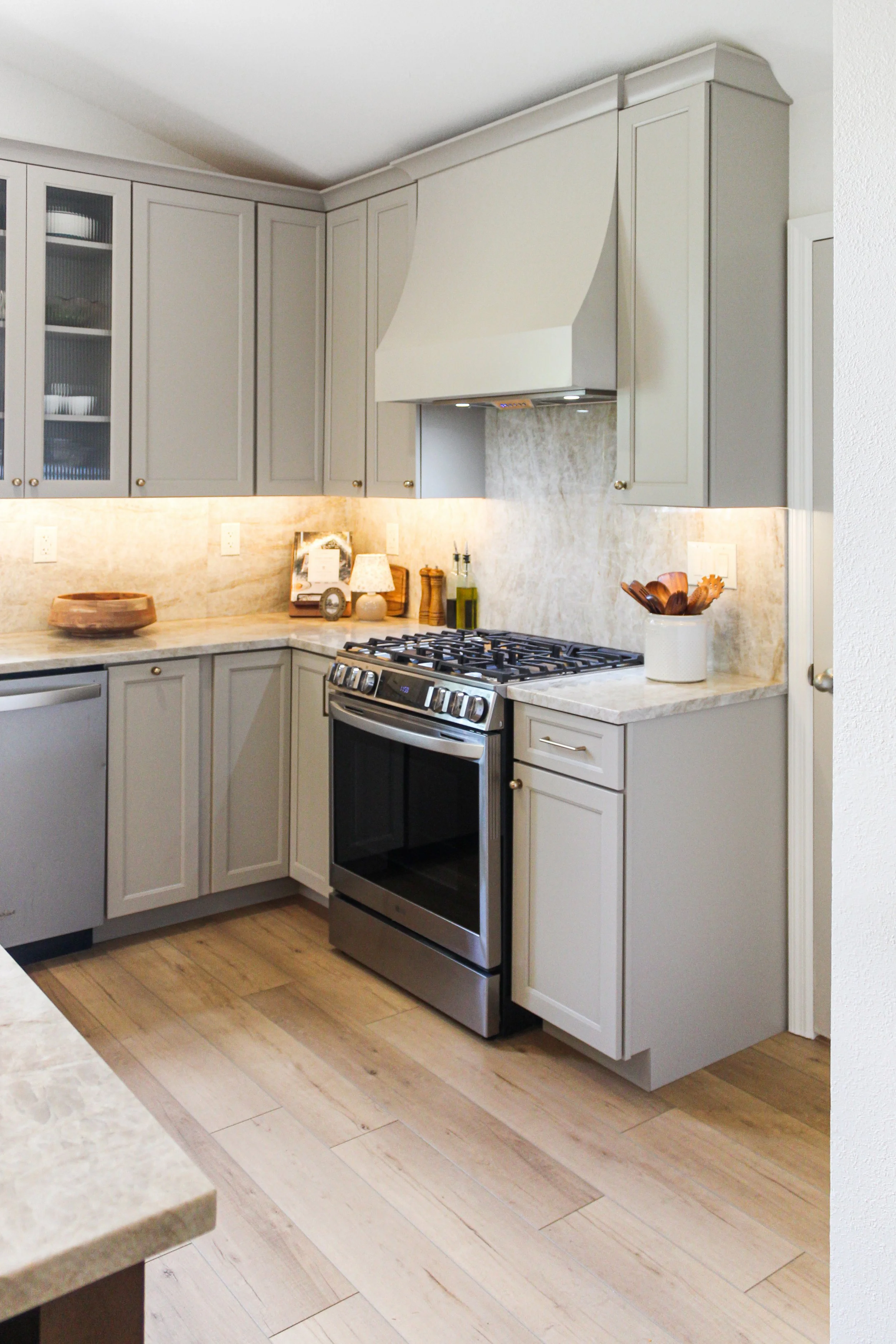 A modern kitchen with beige cabinets, a stainless steel gas stove, a beige granite countertop, decorative items, and wooden floorboards.