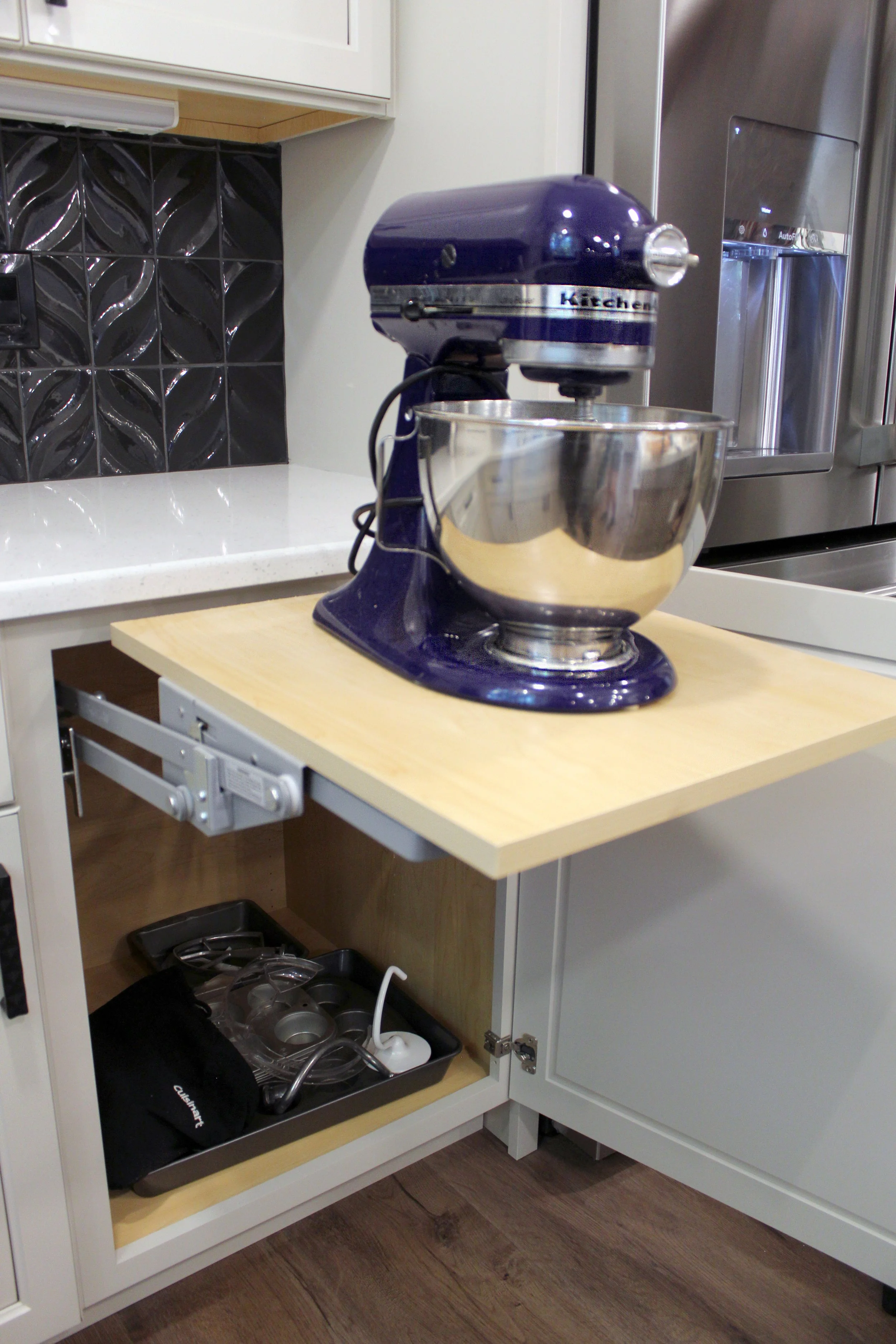 A blue KitchenAid stand mixer with a stainless steel mixing bowl on a wooden counter, next to a refrigerator and black tiled backsplash.