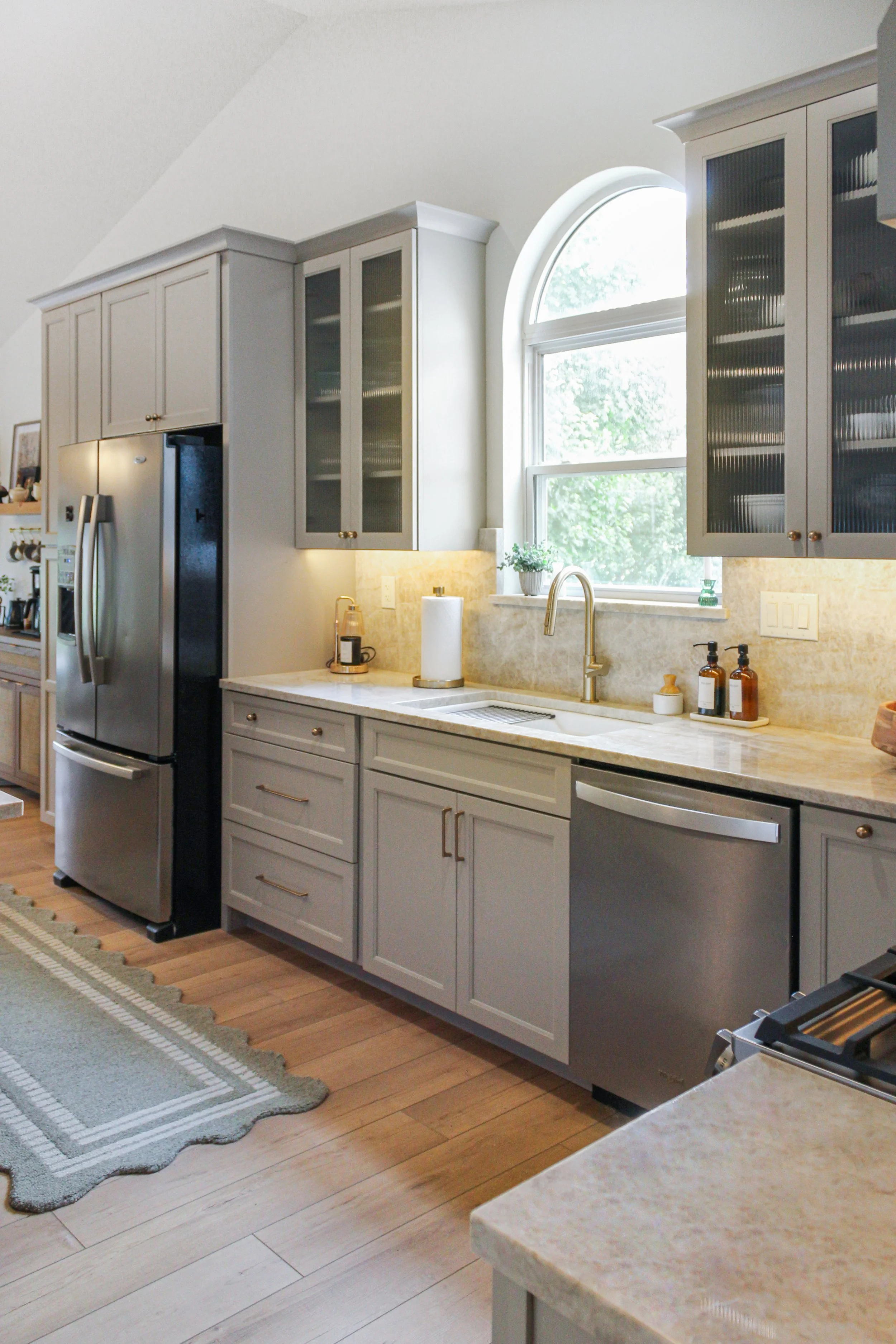Kitchen with gray cabinetry, a large arched window, stainless steel refrigerator and dishwasher, and wood flooring.