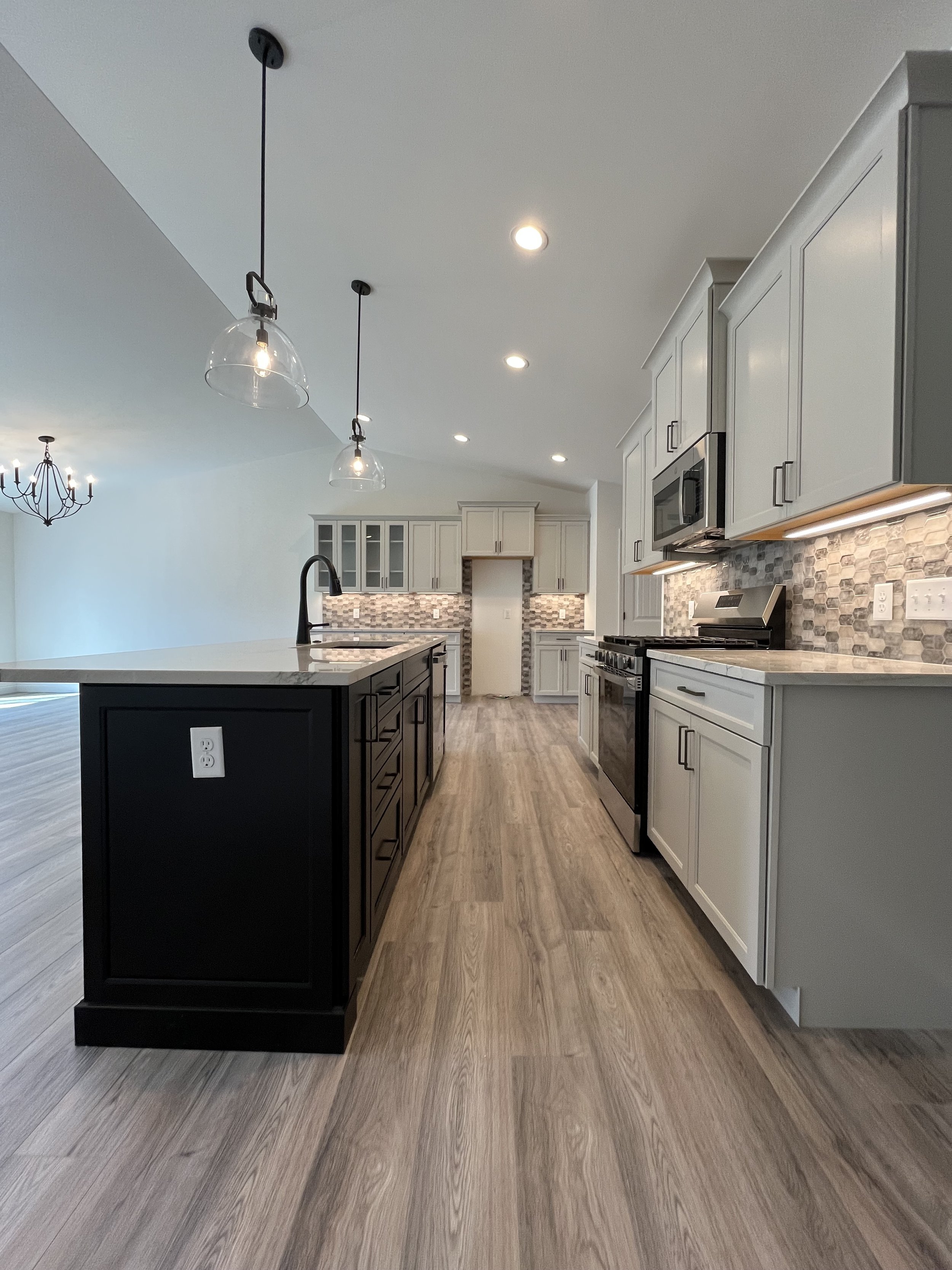 Modern kitchen with gray cabinets, white marble countertops, a black kitchen island, and a patterned backsplash. Pendant lights hang above the island, and hardwood floors are visible throughout.