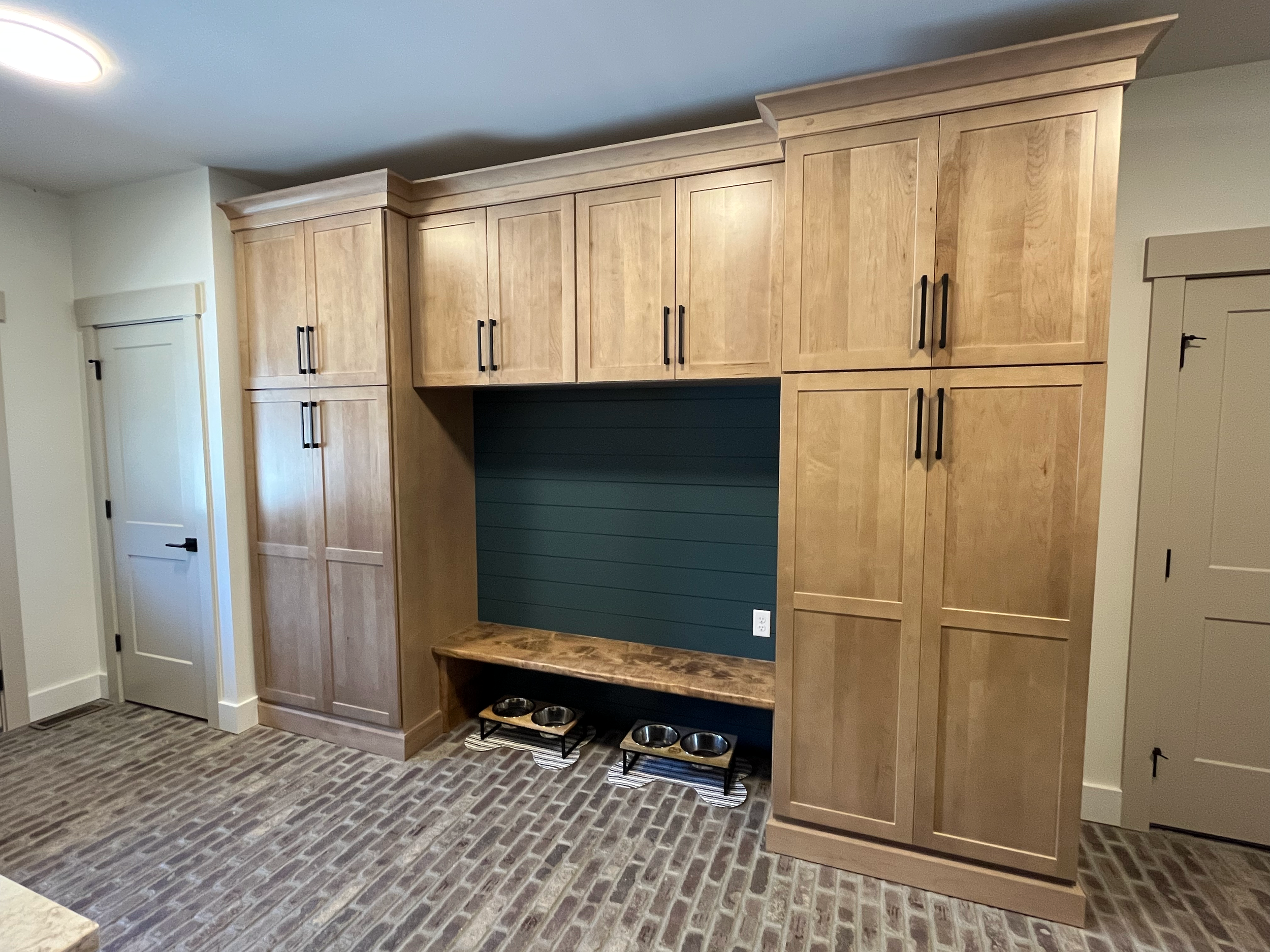 A built-in wooden cabinet with multiple doors and black handles, set against a blue-green painted wall in a room with brick flooring and a power outlet visible at the bottom.