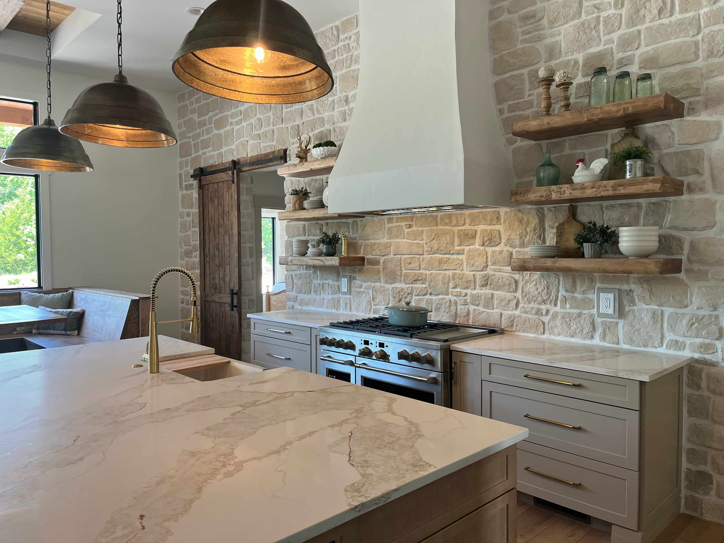 A modern kitchen with a marble countertop, a stainless steel stove, open wooden shelves with decorative items, and distressed white brick walls, illuminated by hanging industrial-style pendant lights.