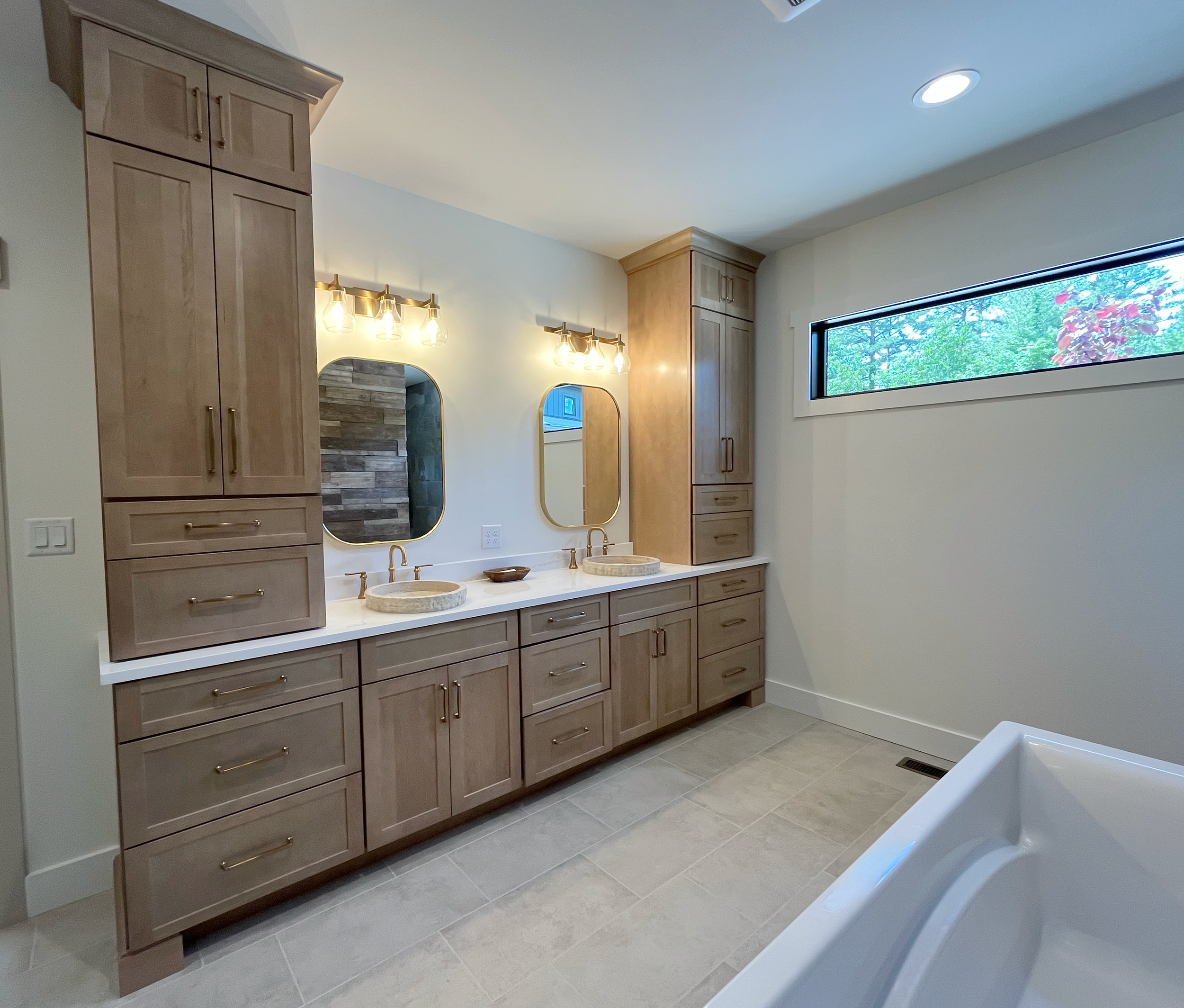Bathroom with wooden cabinets, two mirrors, and a window showing trees and red flowers outside.