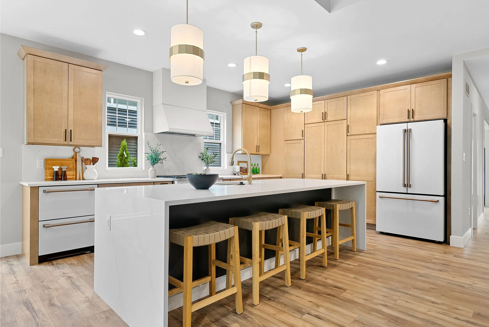 Modern kitchen with white island, wooden cabinets, and stainless steel refrigerator, decorated with pendant lights, plants, and kitchen accessories.