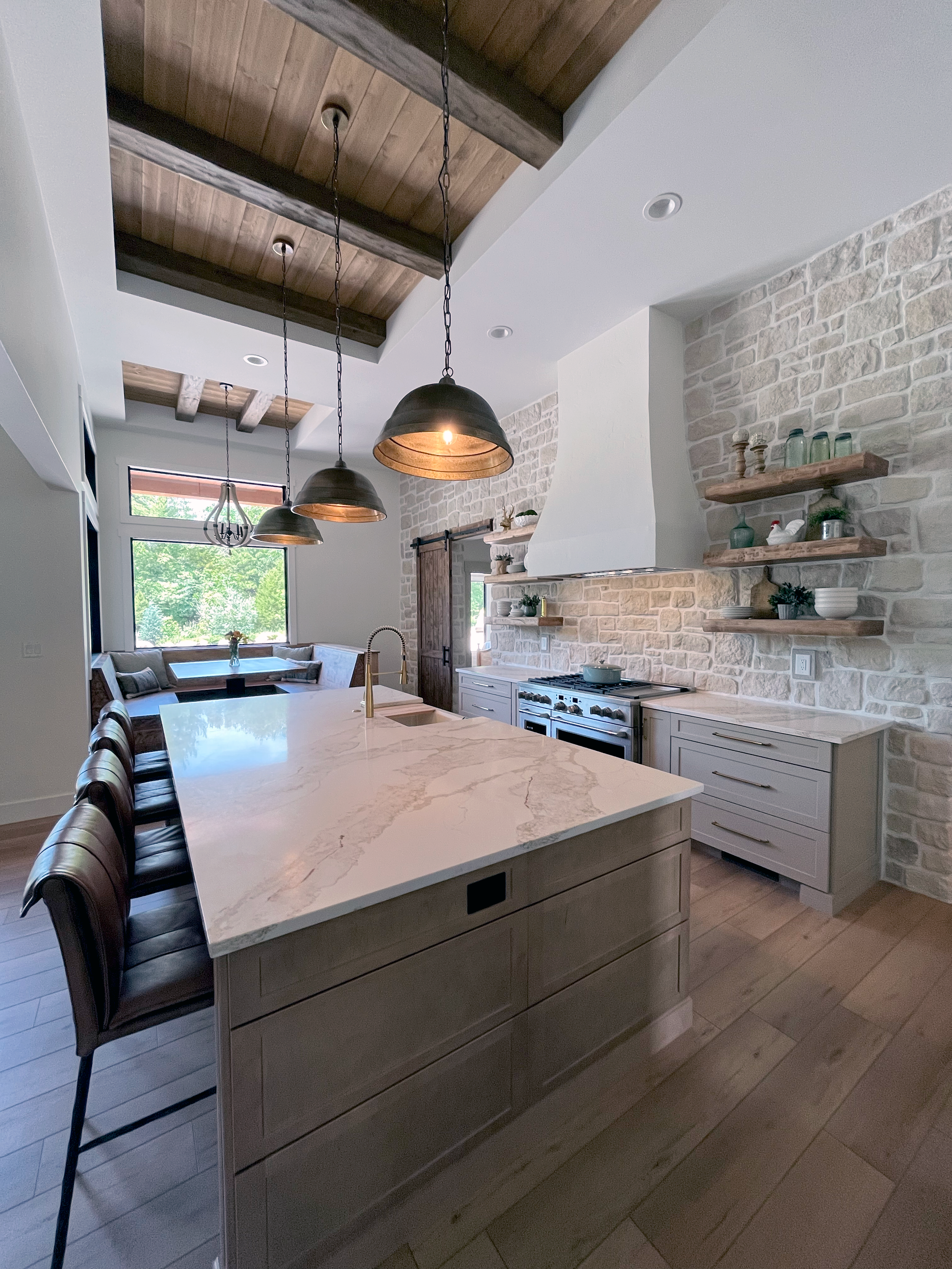 Modern kitchen with a large island with a marble countertop, pendant lights hanging above, a stove against a brick wall, and open wooden shelves with decorative items.