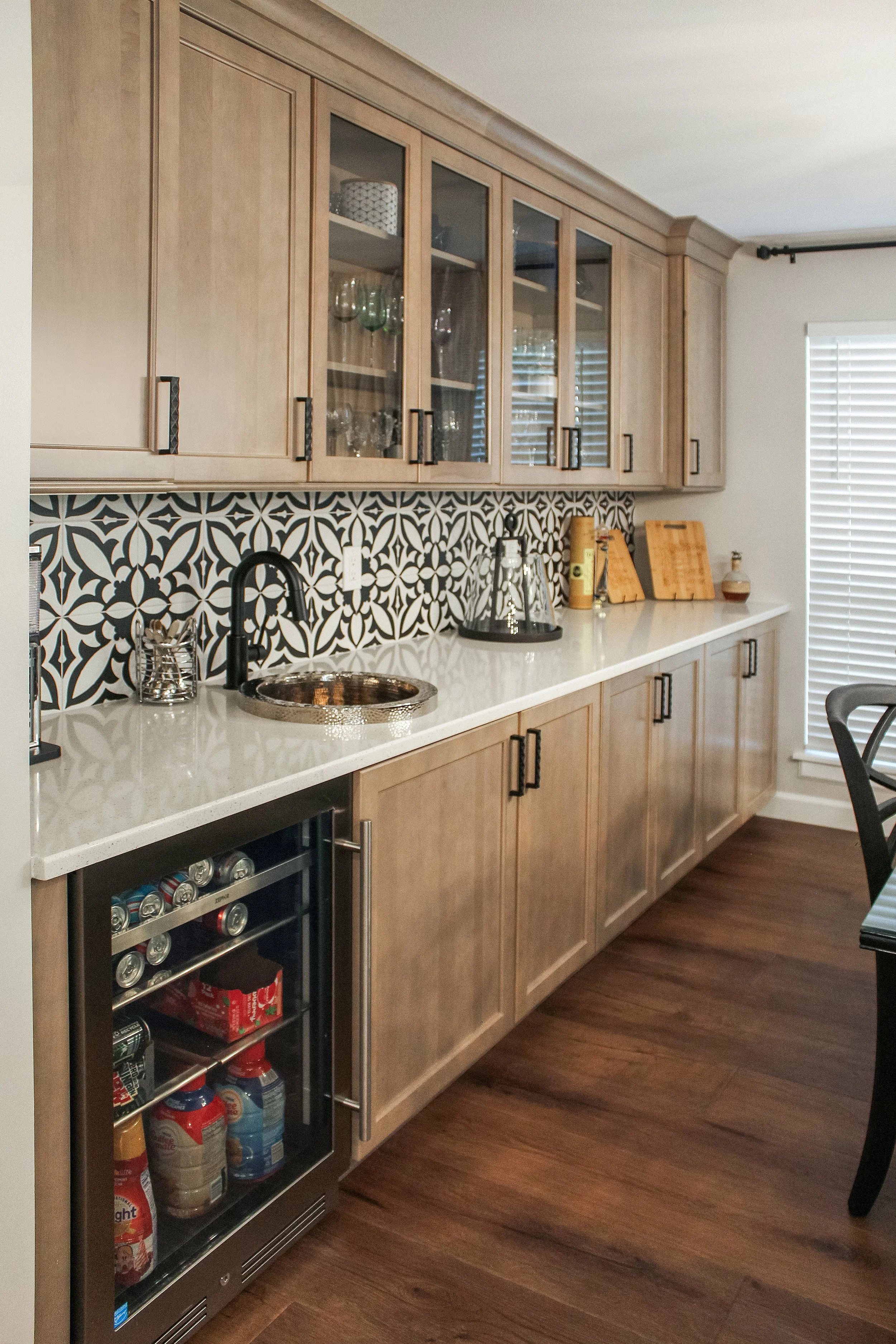 Kitchen with wooden cabinets, patterned black and white tile backsplash, small sink, and mini fridge stocked with cans and bottles.
