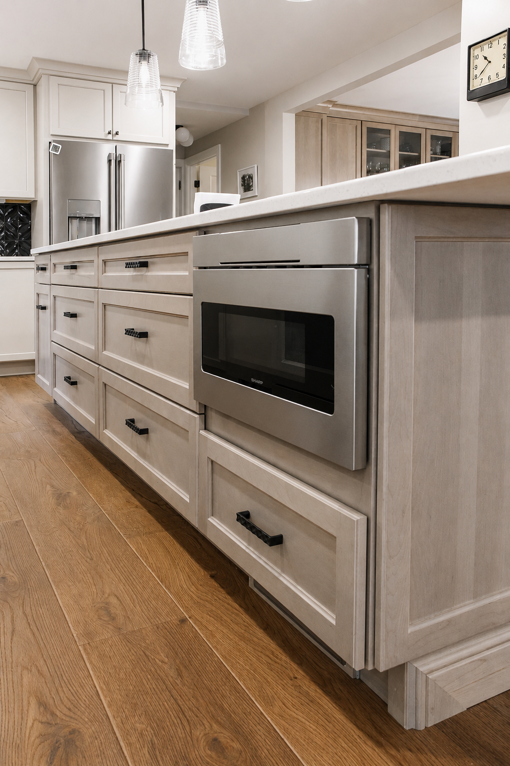 Kitchen island with drawers and an integrated microwave oven, with a stainless steel refrigerator in the background and pendant lights hanging from the ceiling.