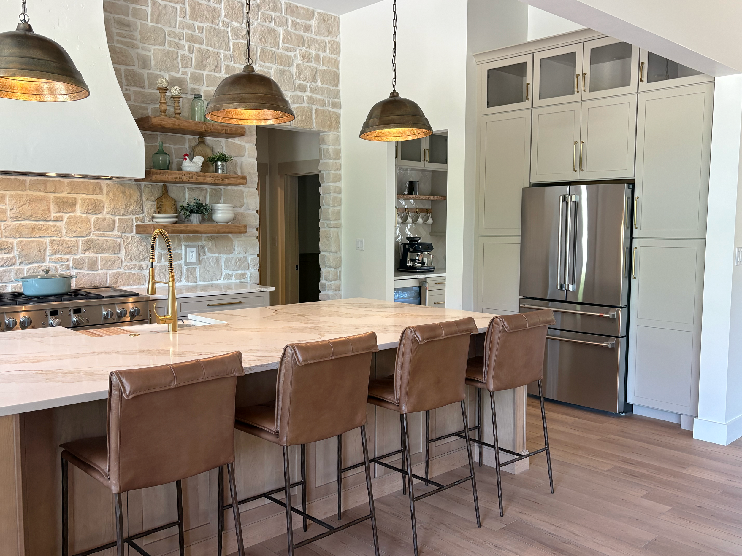 Modern kitchen with a stone accent wall, hanging bronze pendant lights, a marble countertop island with brown leather bar stools, and integrated white cabinets with gold handles surrounding stainless steel appliances.