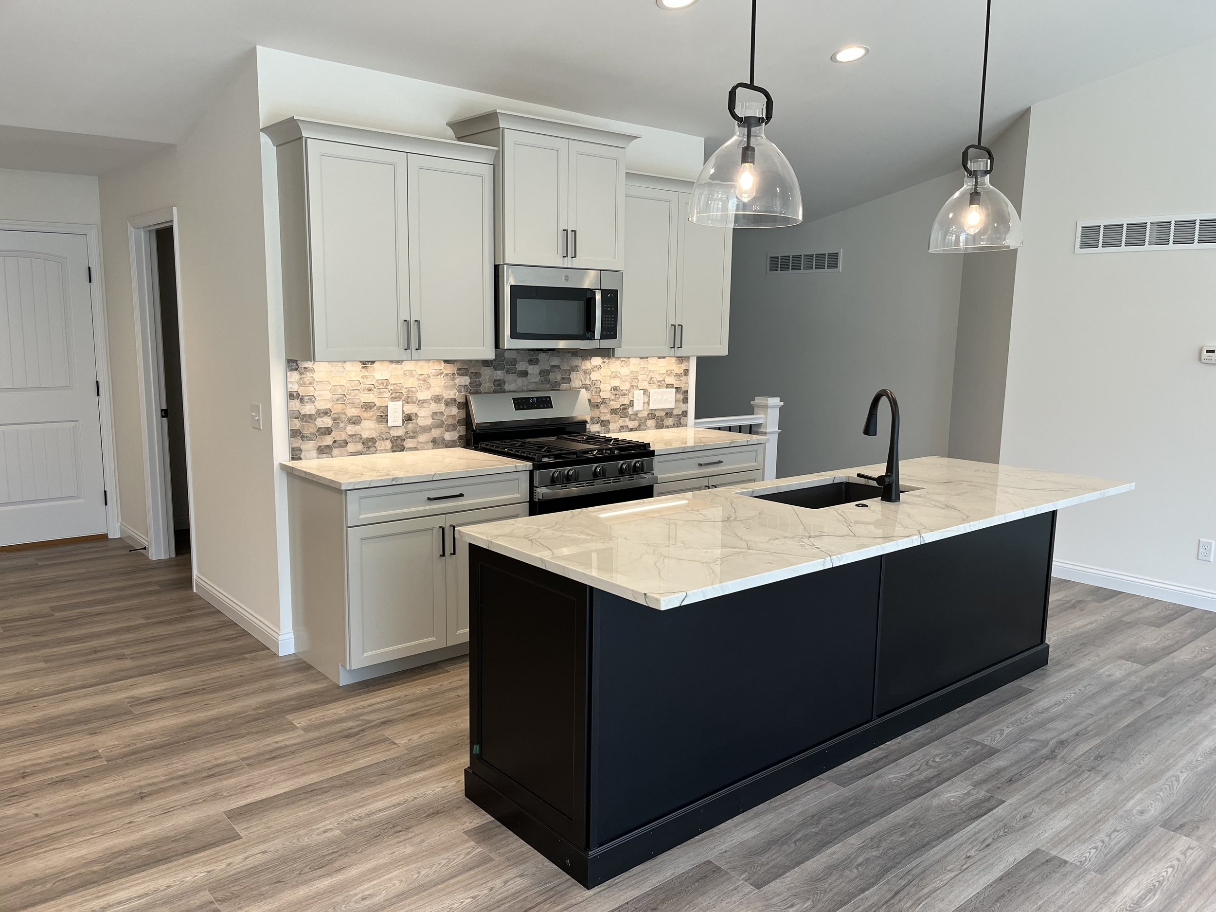 Modern kitchen with white cabinetry, a marble island with a black base, a stainless steel microwave above a stove, pendant lights hanging from the ceiling, wood-look flooring, and a tiled backsplash.