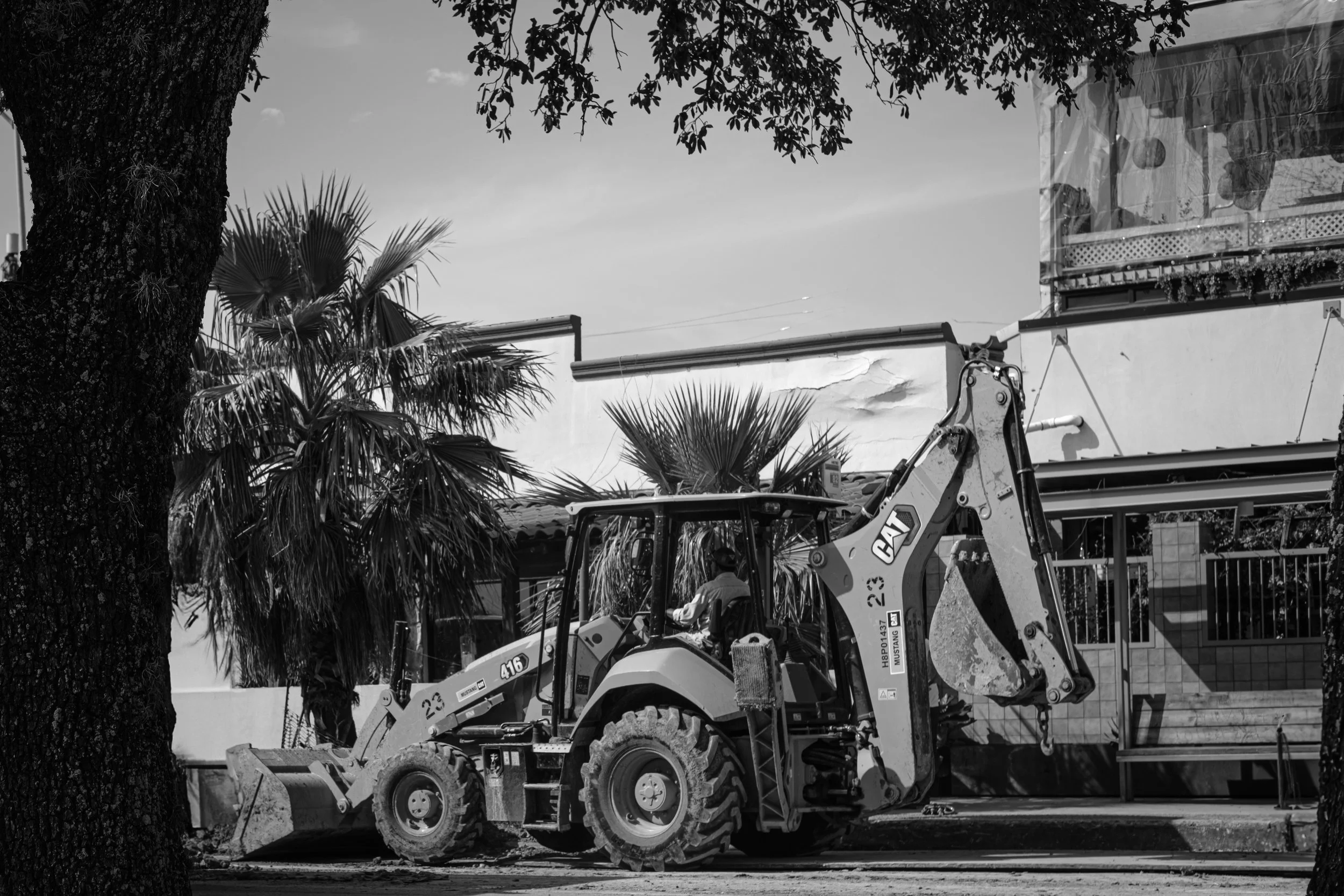 A backhoe loader parked on a street in front of a building with palm trees nearby.