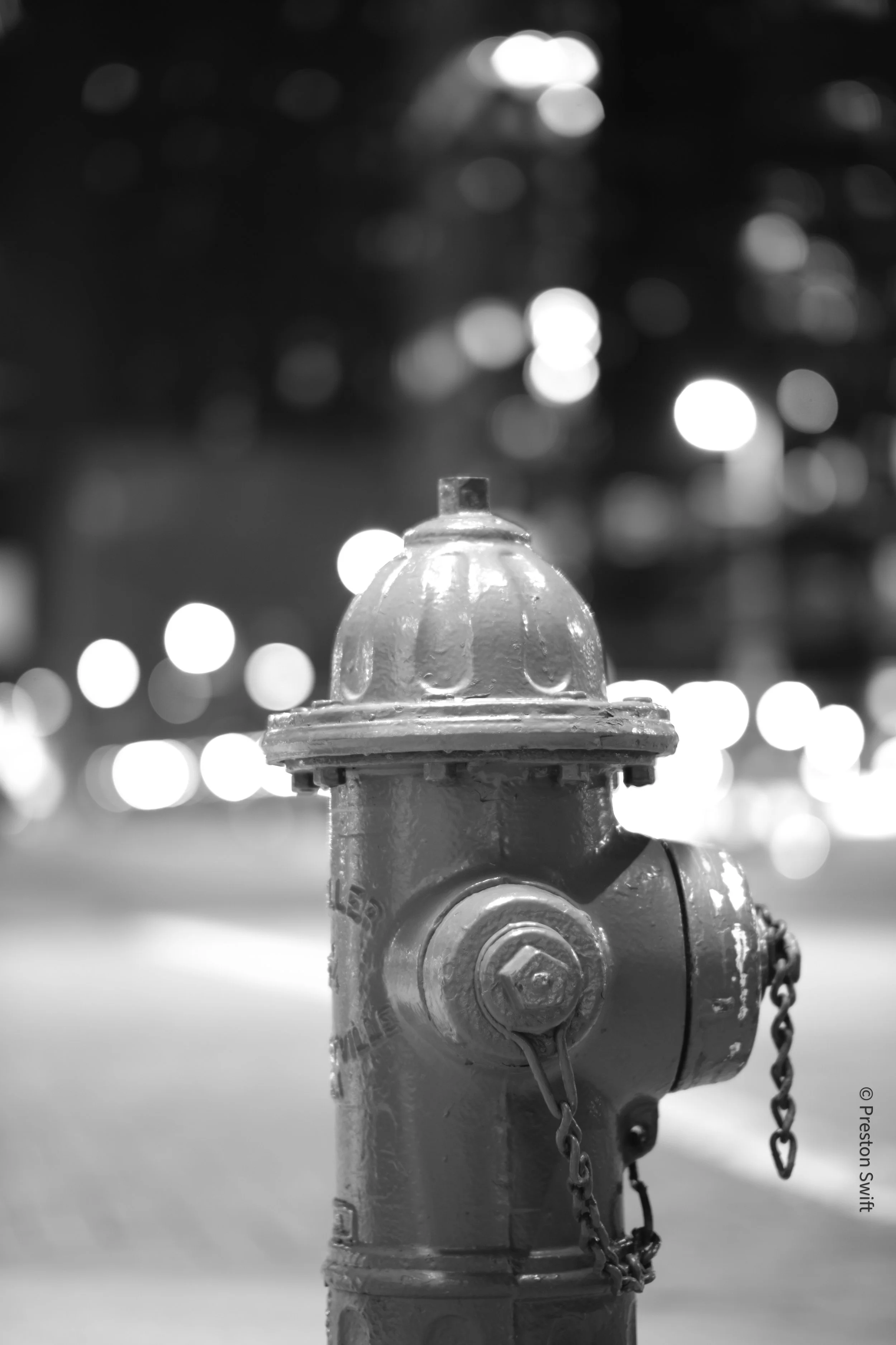 A black and white photograph of a fire hydrant on a city street at night, with blurred city lights in the background.