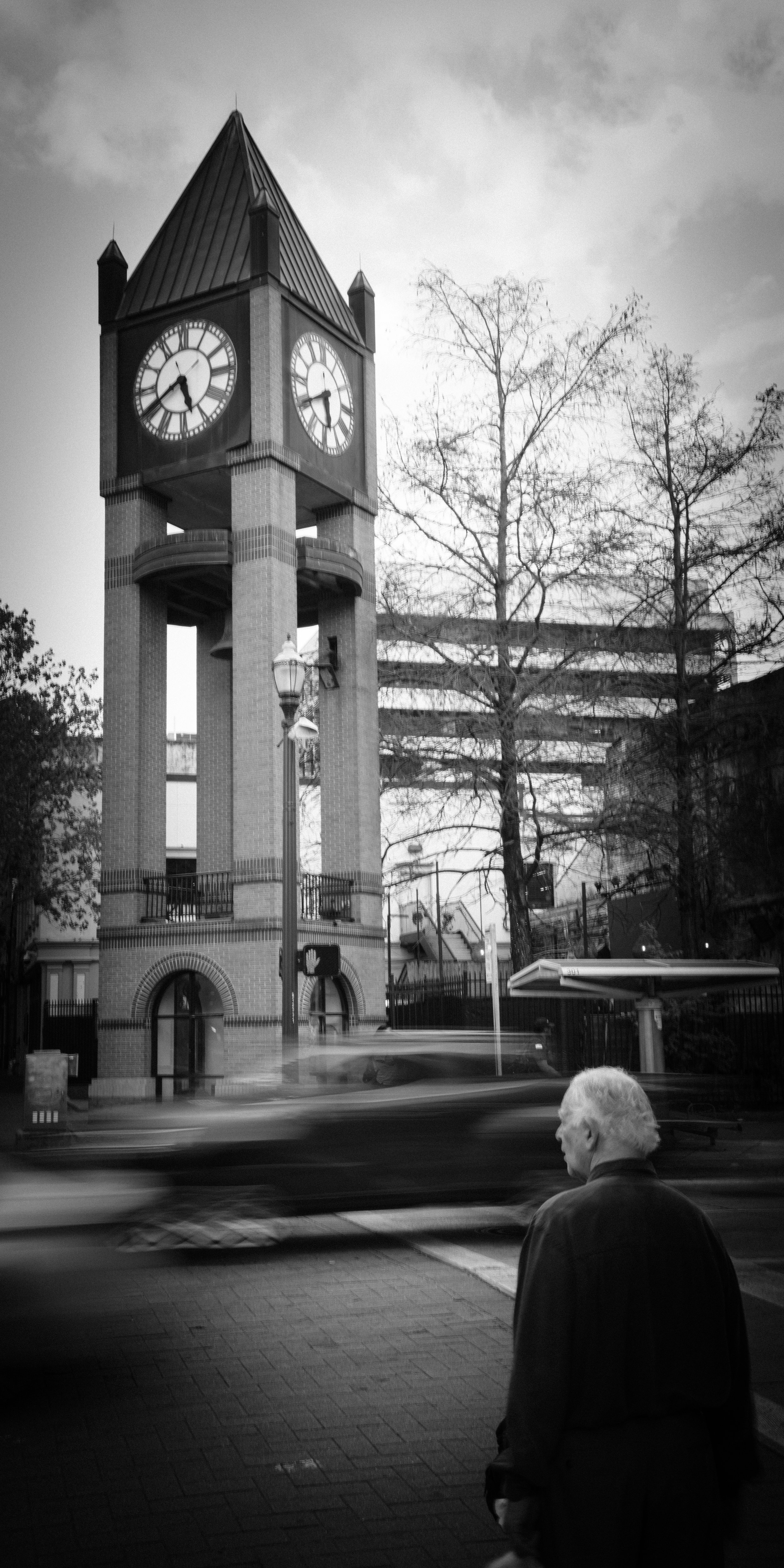 Black and white photo of a clock tower with a man standing nearby on a city street, blurred cars passing by, and leafless trees in the background.