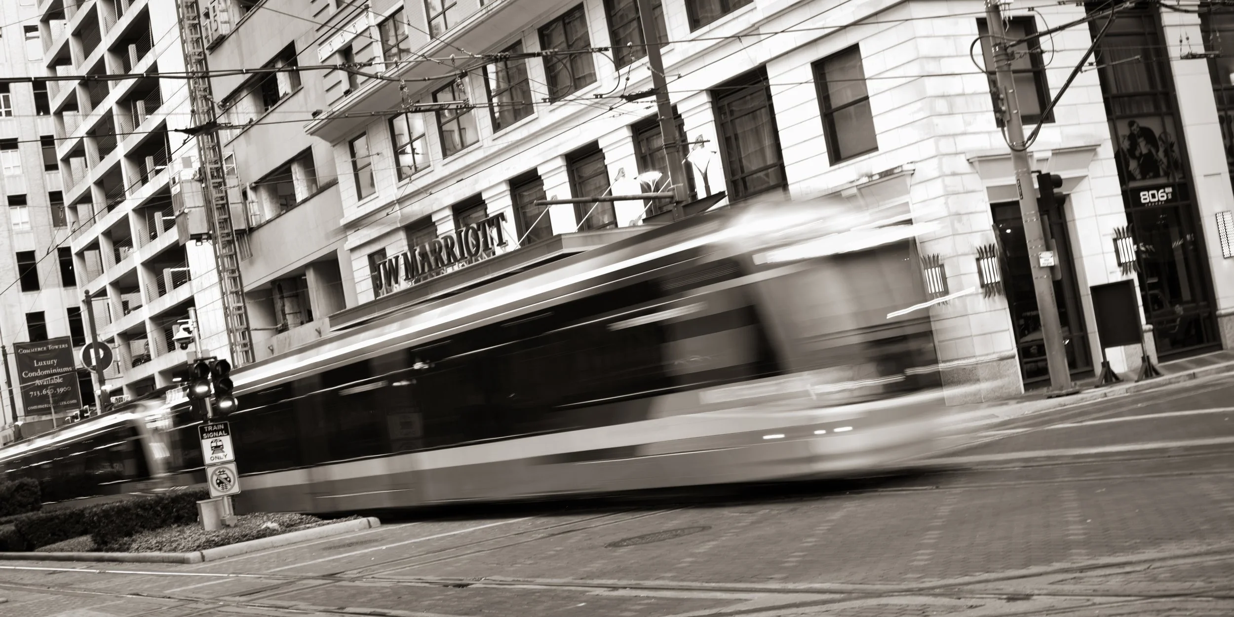 Black and white photo of a moving tram in an urban city street with buildings and street signs.