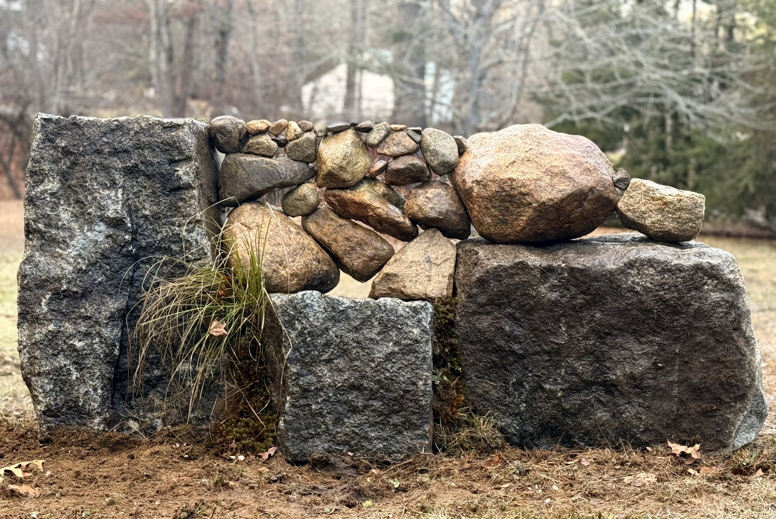 A dry stone wall built with large and small rocks, with some grass and moss growing at the base, outdoors during autumn or winter with trees in the background.