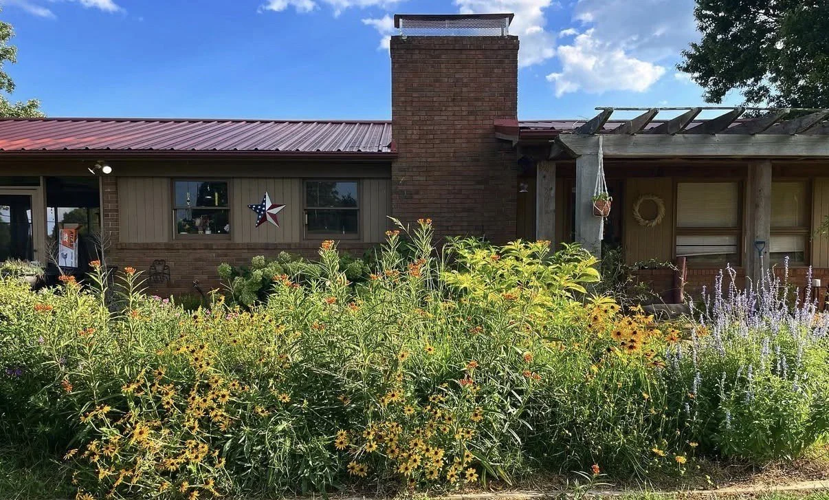 A house with a brick chimney, surrounded by a colorful garden with yellow, orange, and purple flowers under a partly cloudy sky.