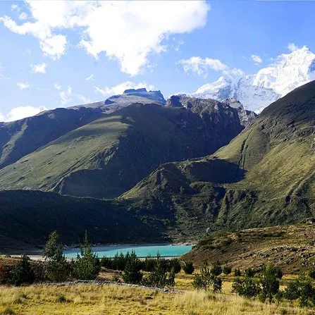Paisaje montañoso con un lago turquesa, colinas verdes y montañas cubiertas de nieve al fondo bajo un cielo soleado con algunas nubes.